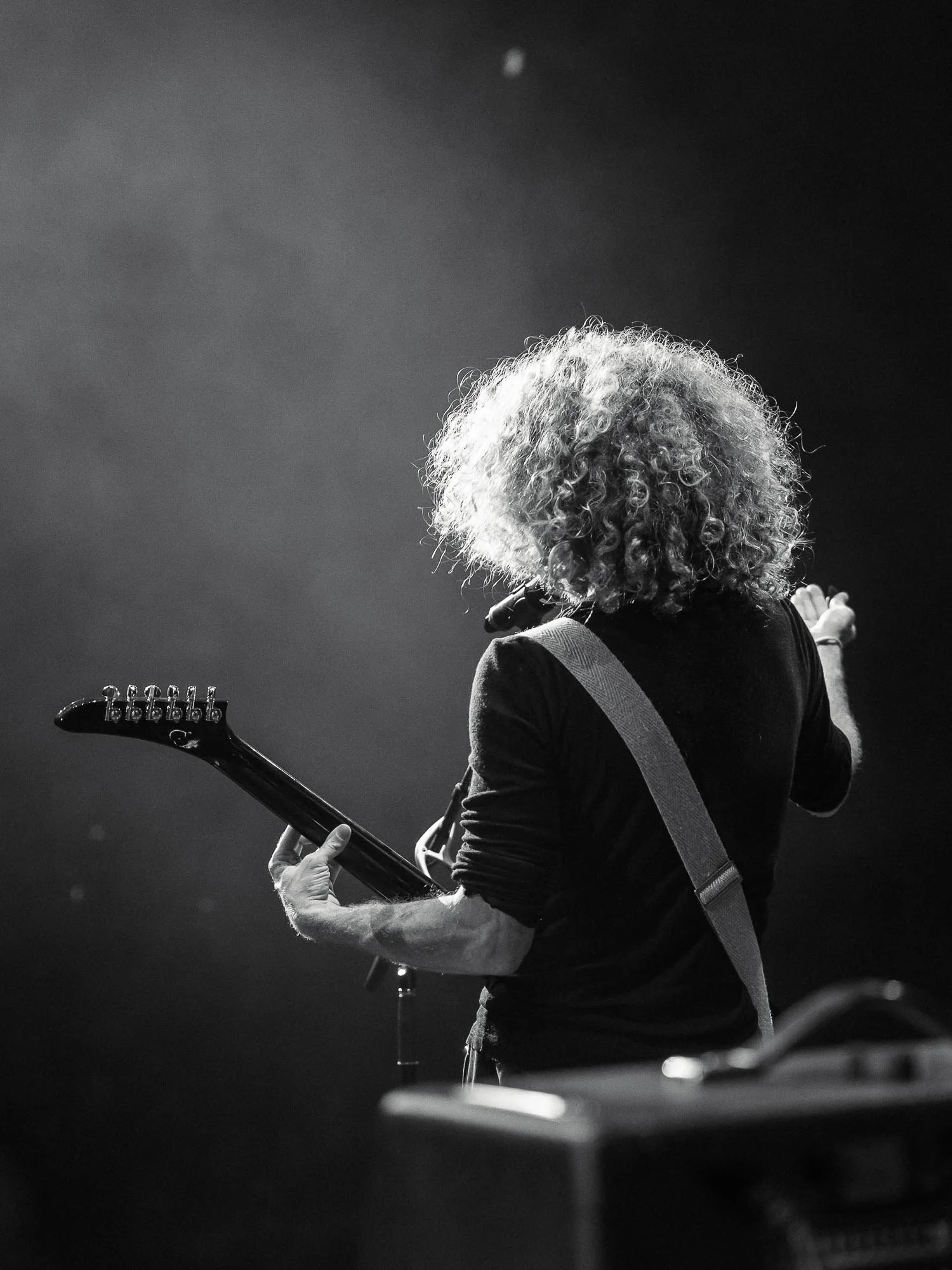 A black-and-white photo of a Glenn with curly hair playing an electric guitar on stage, facing away from the camera, with a spotlight shining down and a dark background.