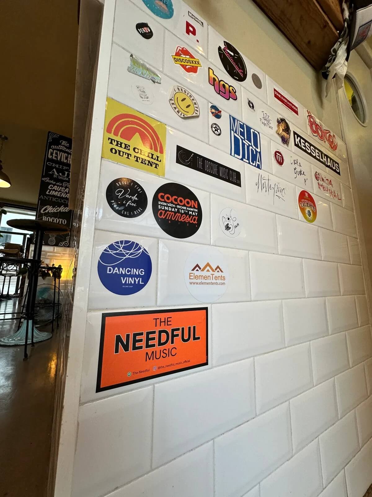 A wall decorated with various music and event stickers, including logos and signatures, in a room with a glimpse of dining tables and chairs.