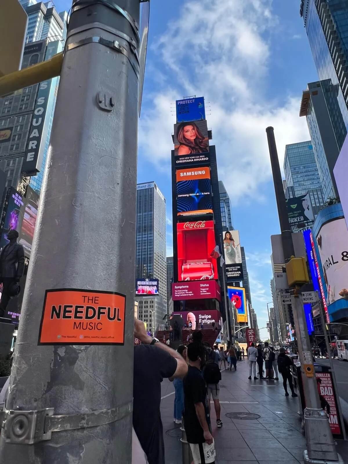 Time Square in New York City with illuminated digital billboards, people walking on the sidewalk, and a gray pole with an orange sticker reading "The Needful Music."