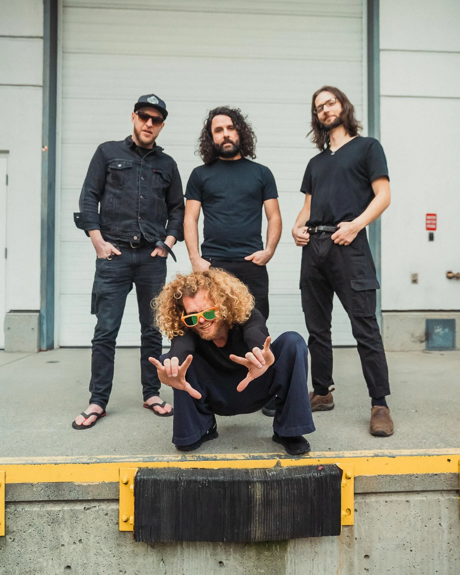 The Needful posing in front of a white garage door; one man sitting on the ground with curly blonde hair and sunglasses, three men standing, dressed casually with one in flip-flops.