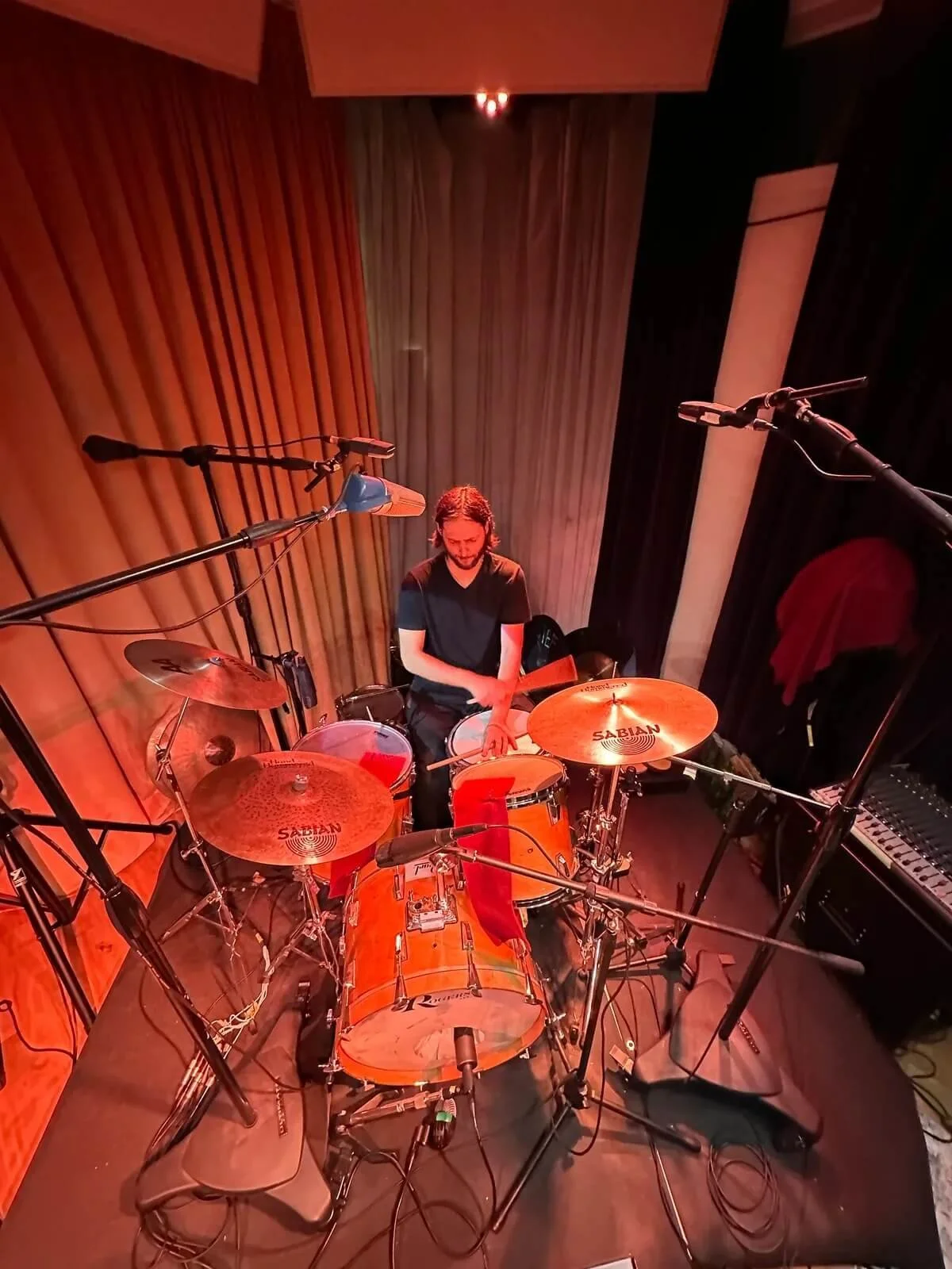 The Needful drummer with shoulder-length hair and a beard playing a drum kit on a stage with wooden panel walls, surrounded by microphones and audio equipment.