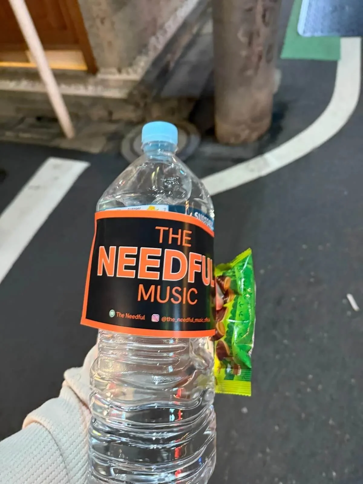 A water bottle with a black and orange label reading 'The Needful Music' being held outdoors on a street. The bottle has a blue cap and is held by a person wearing a white sweater, with a snack attached to the bottle. The street has a curb, a tree tr