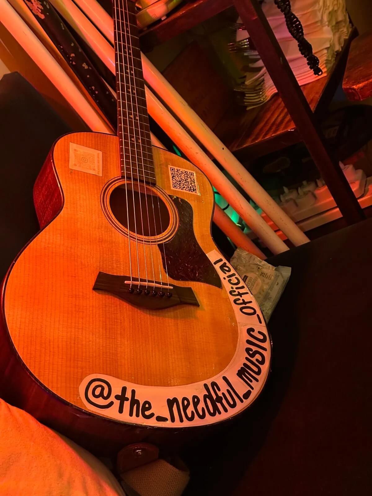 An acoustic guitar with a sticker that reads '@the_needful_musical_official' resting on a black chair in a dimly lit room with wooden shelves and dishes in the background.