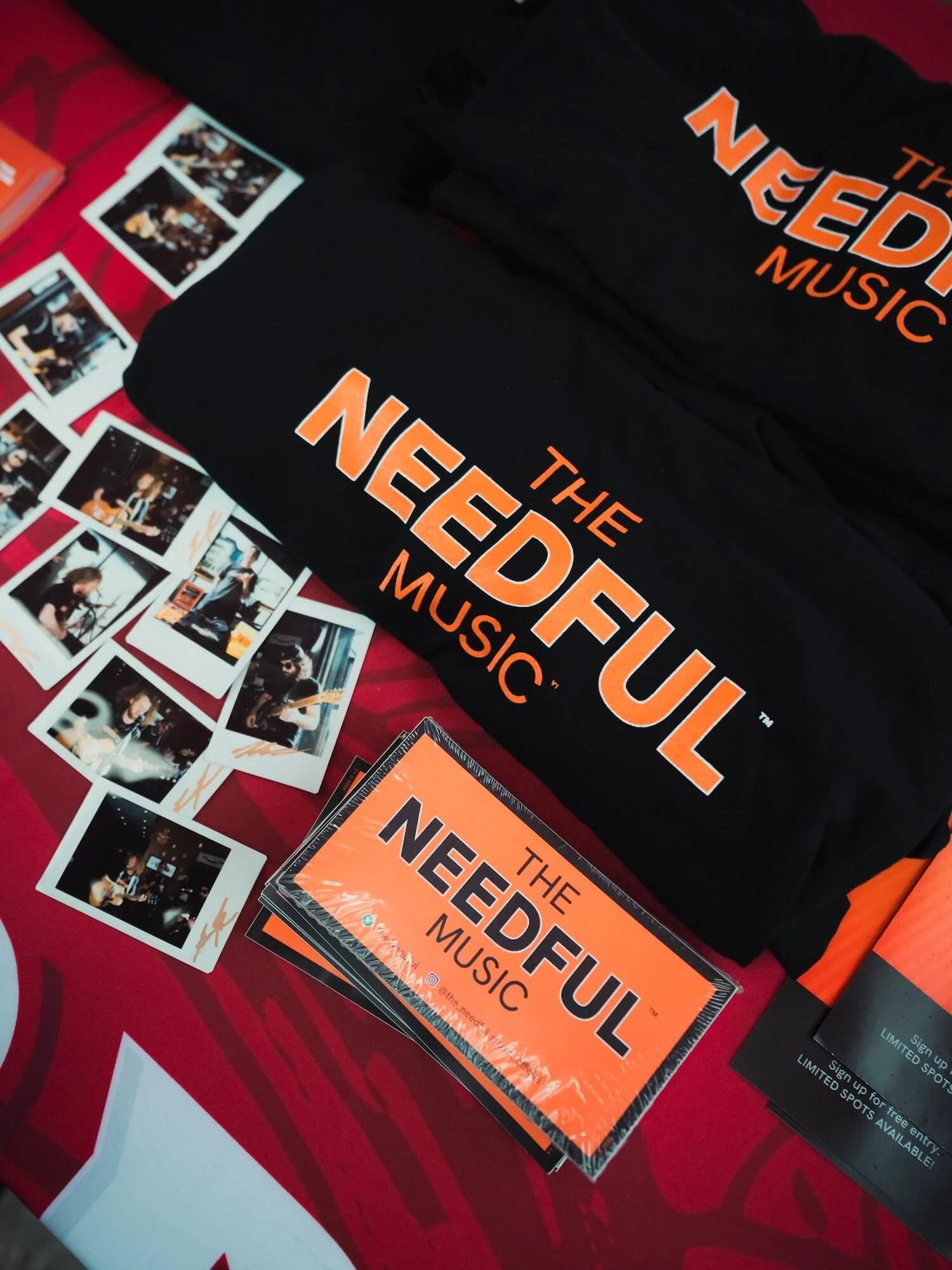 Black T-shirts and orange printed cards with the words "The Needful Music" displayed on a red table with many photographs of musicians.