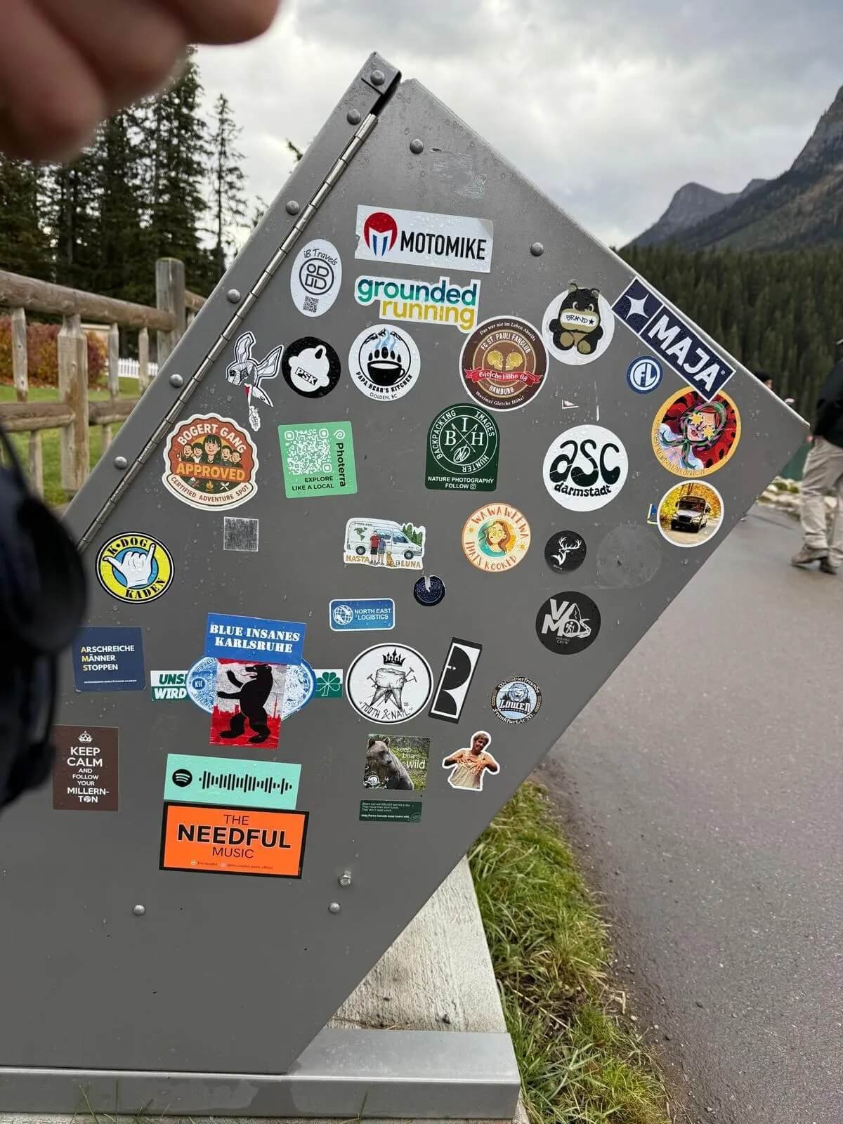 A metal signboard covered with various colorful stickers in an outdoor setting with trees and mountains in the background.