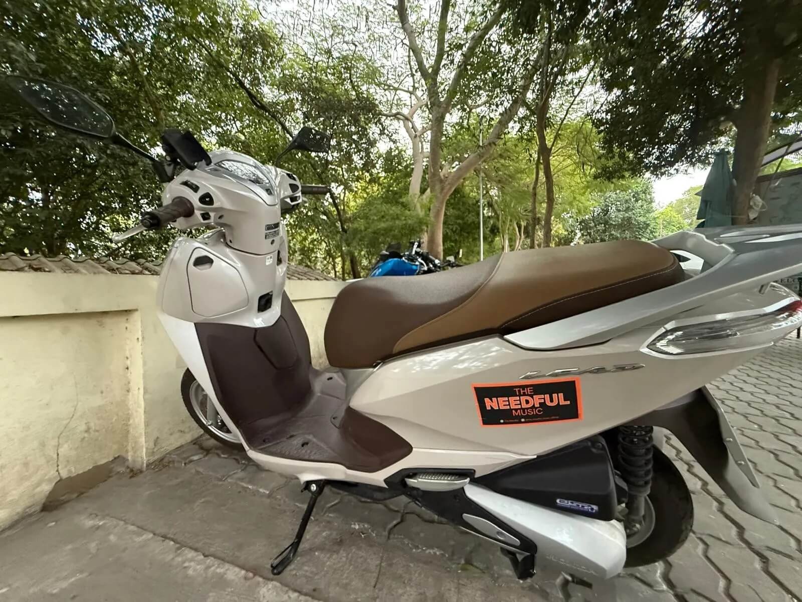 A white and brown scooter parked on the sidewalk next to a beige concrete wall, with trees and a blue motorcycle in the background.