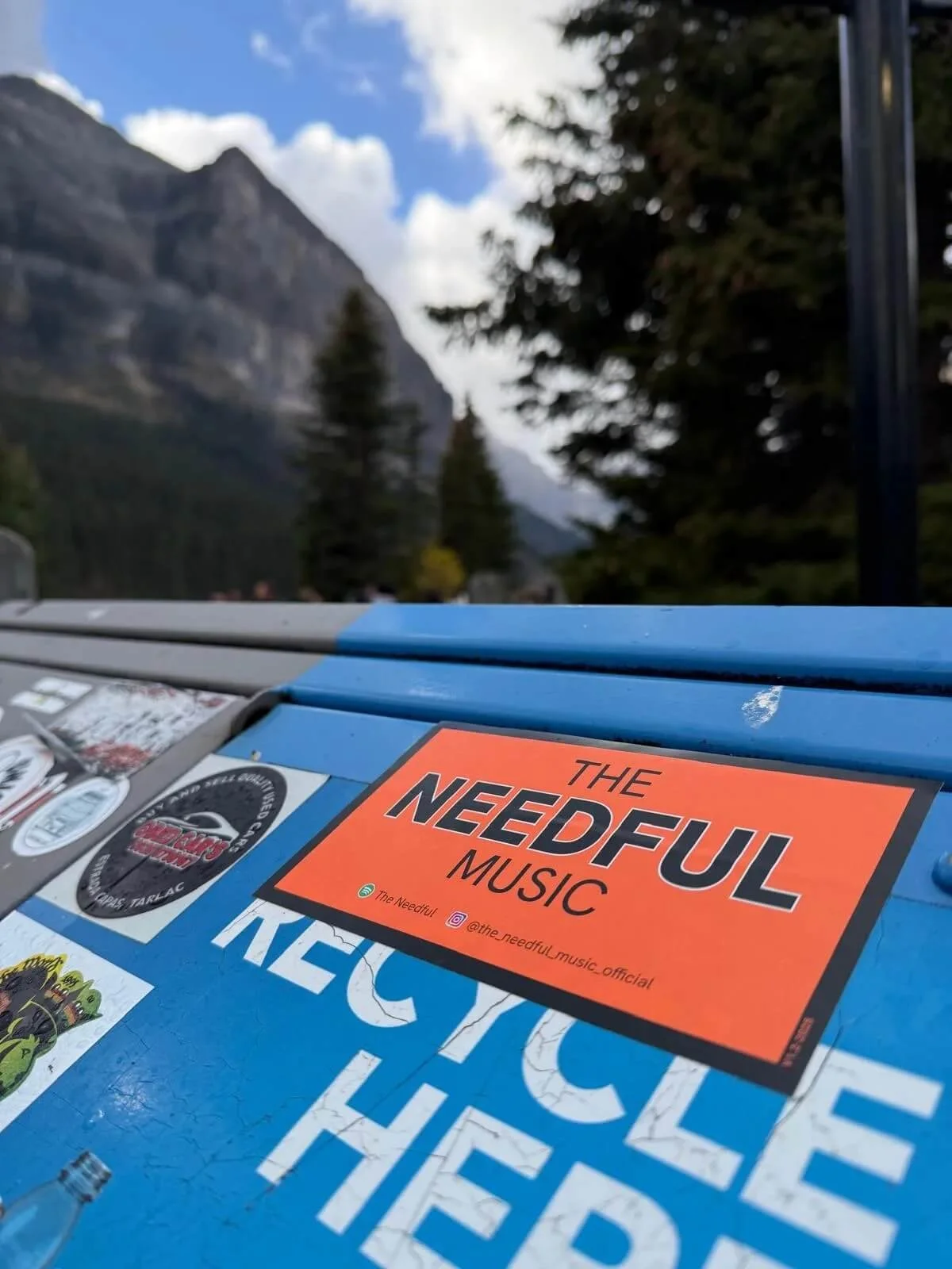 A close-up of a blue surface with stickers, including an orange sticker that reads 'The Needful Music'. In the background, a mountainous landscape with trees and partly cloudy sky.