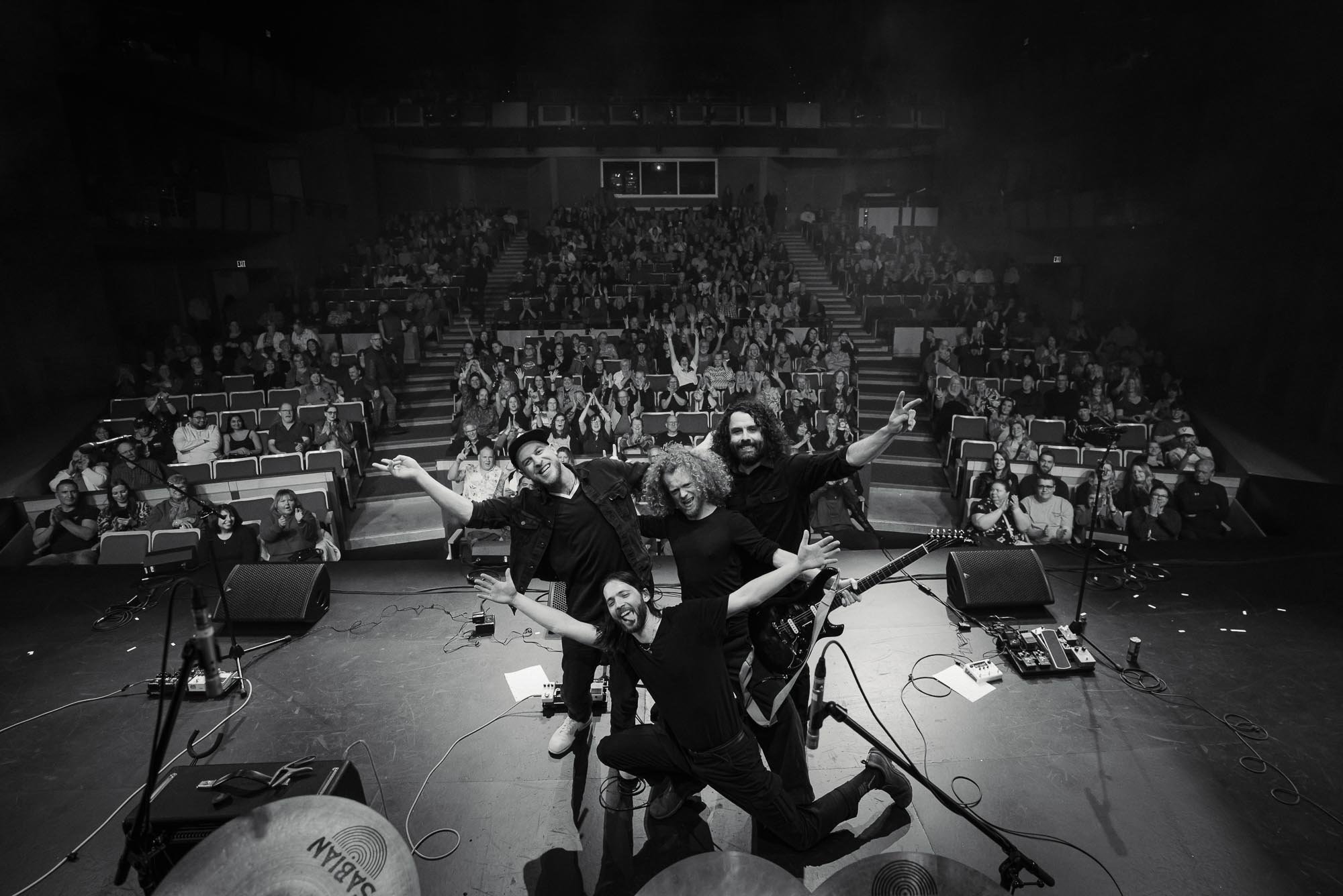 Three musicians from The Needful on stage taking a group selfie with a large audience in the background, in Duncan, BC. The venue is a theatre or concert hall with seats filled with fans.