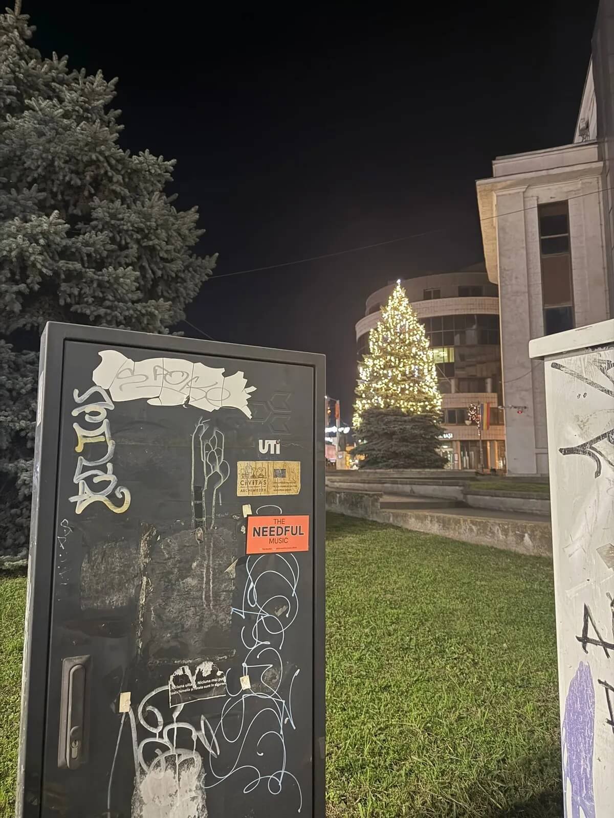 Night scene featuring a decorated Christmas tree with white lights, a large building, a tree, and a black phone booth with graffiti and stickers.