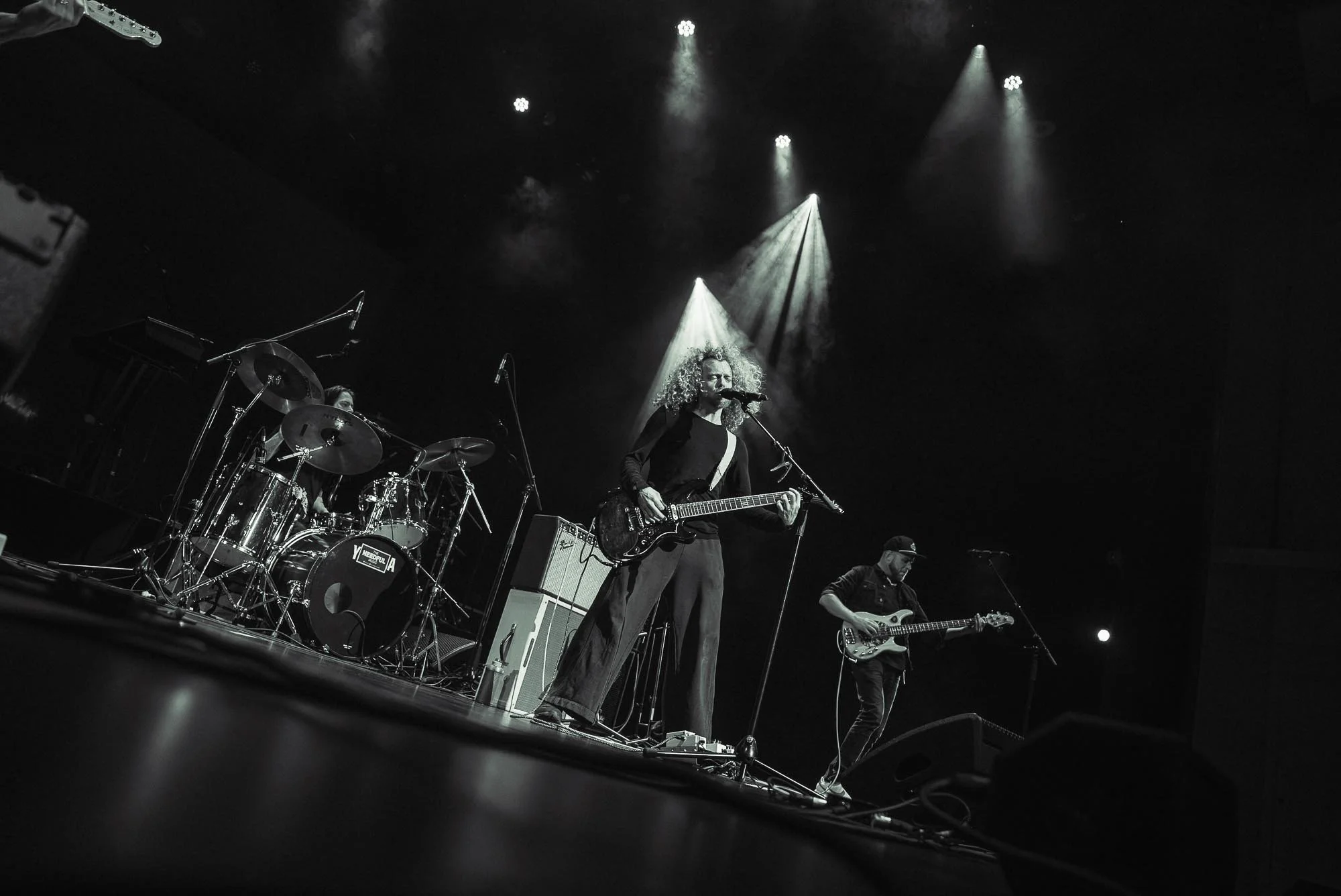 A live musical performance on stage with a band. A woman with curly hair singing into a microphone and playing an electric guitar is center stage, surrounded by other musicians, including a drummer and guitarist. Stage lights shine down, creating a d