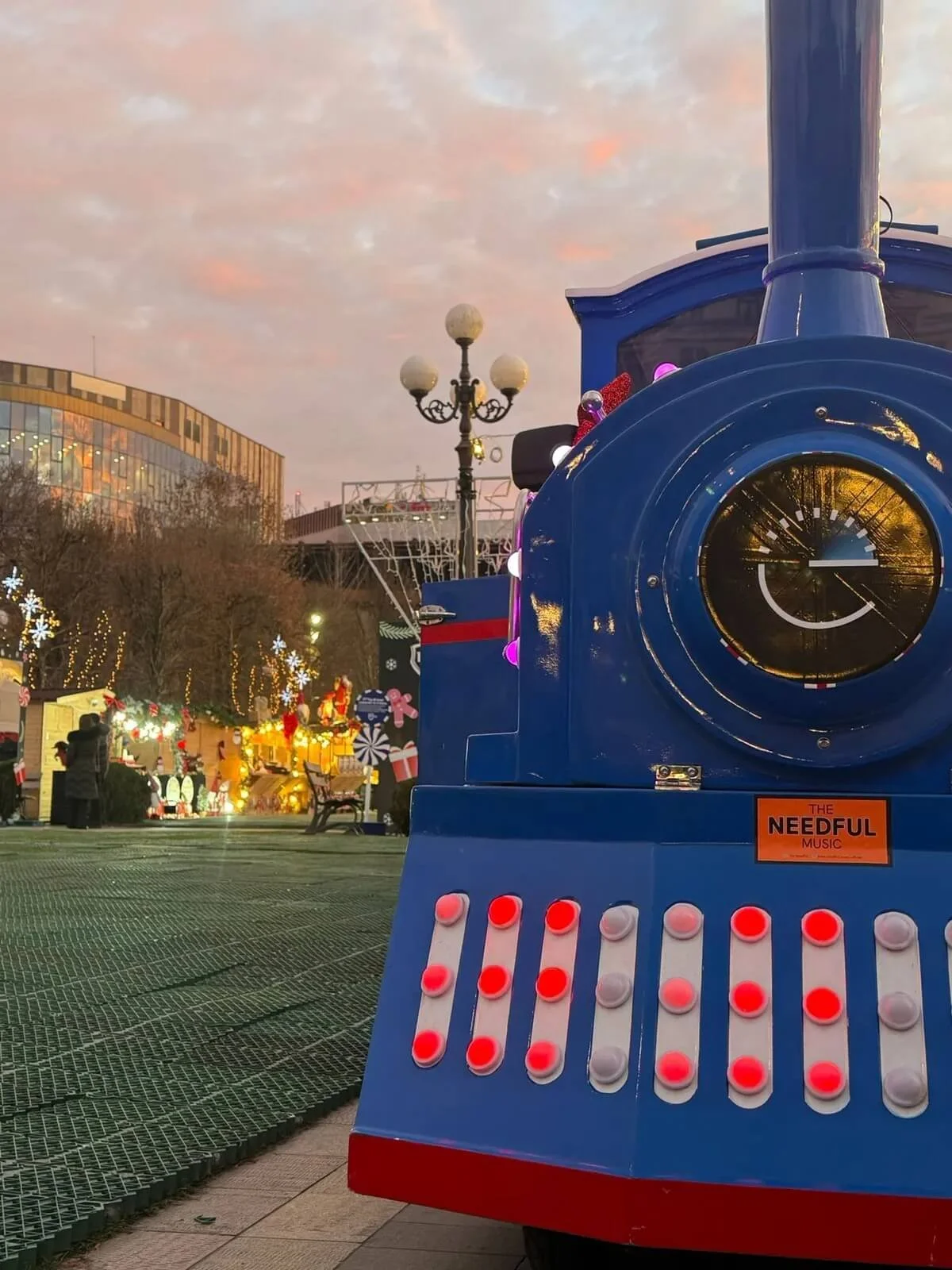 Close-up of a blue train decoration with colorful lights in an outdoor festive setting during sunset, with people and holiday decorations in the background.