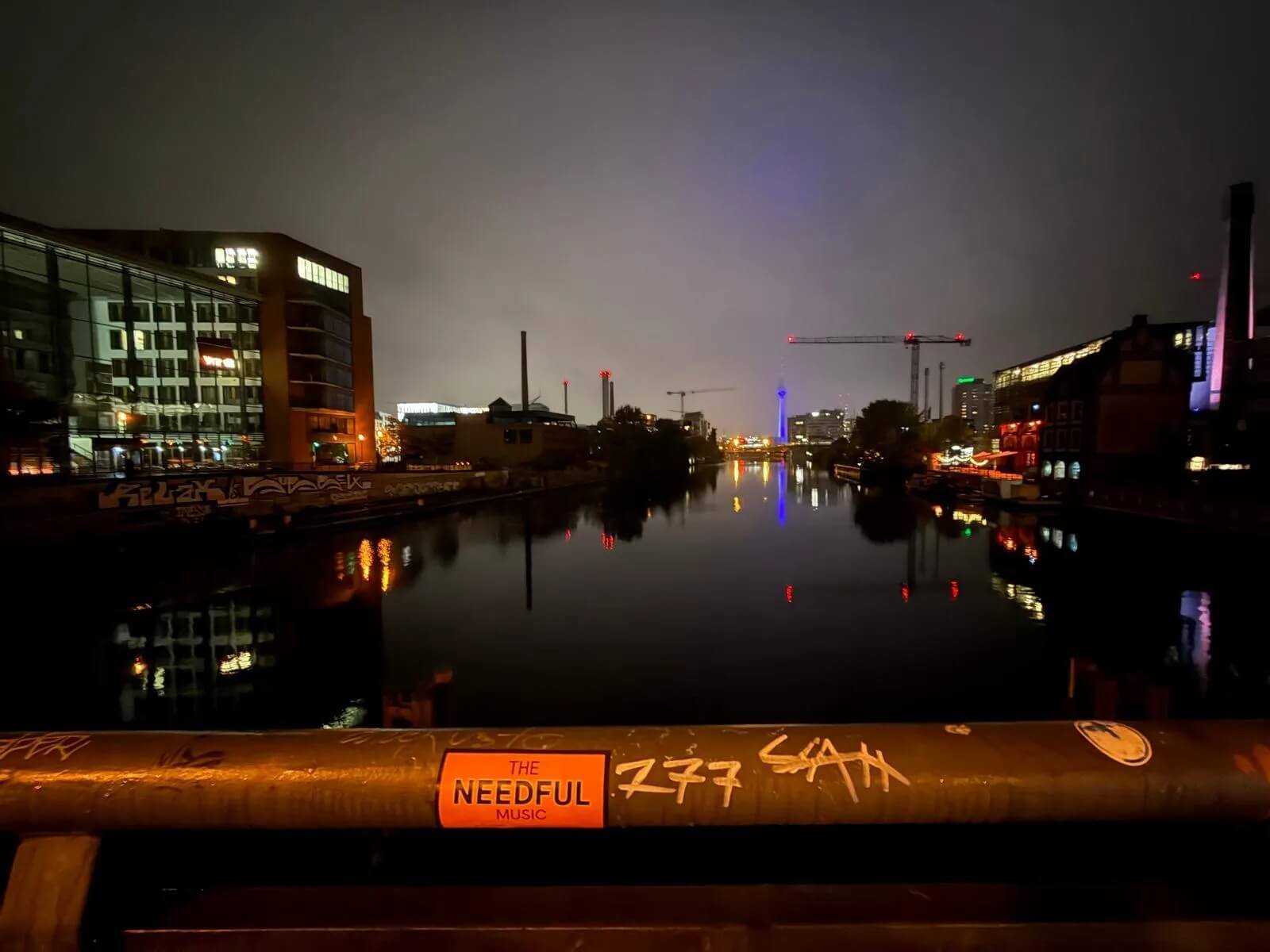 Nighttime cityscape with illuminated buildings and construction cranes reflected on a river, viewed from a bridge with graffiti and an orange sticker that reads 'THE NEEDFUL MUSIC'