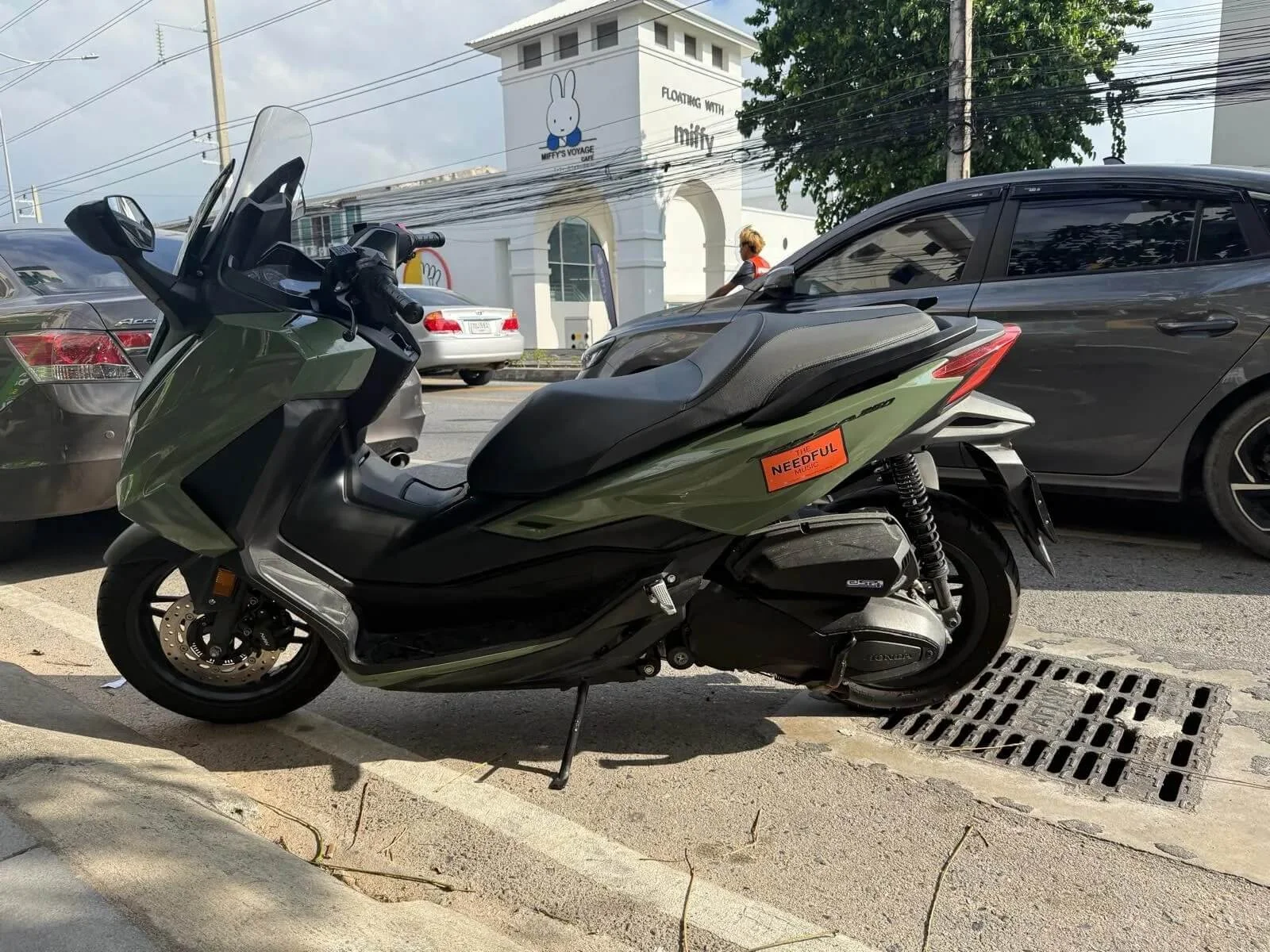 Green and black scooter parked on a sidewalk next to a curb with a drain grate visible and The Needful sticker. Cars are parked behind it on the street, and a person with blonde hair and a red shirt is walking past.