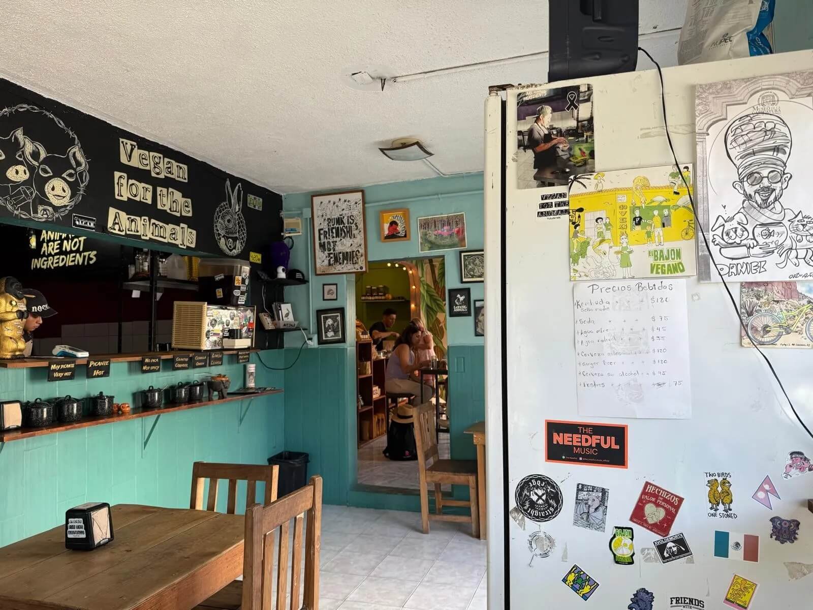 Interior of a vegan restaurant with teal walls, decorated with blackboard art and framed pictures, with a white refrigerator covered in stickers, notes, and drawings, and a doorway leading to a dining area with customers seated at tables.