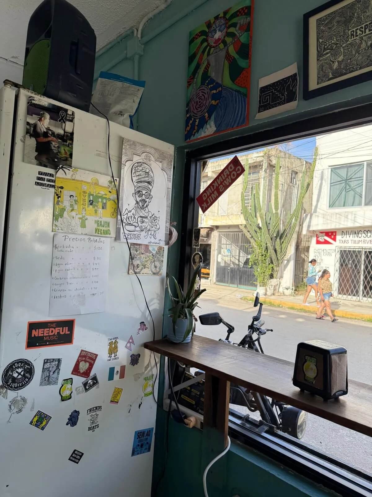 Interior of a small shop with a white refrigerator covered in stickers and drawings, a small wooden table with a potted plant and a vintage radio, and a large window revealing people walking outside on a sunny day.