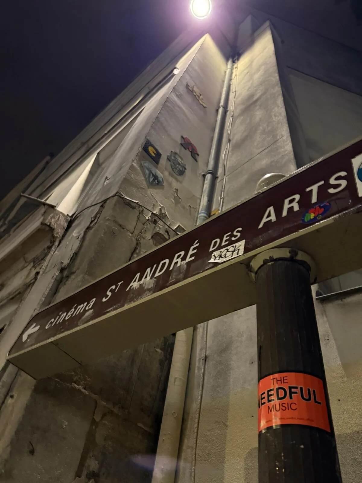 Nighttime street scene featuring a building with a street sign for 'Cinemas St. André des Arts' and stickers on a pole that reads 'The Neeful Music'. The building's wall has some street art or posters, with a bright streetlight illuminating the