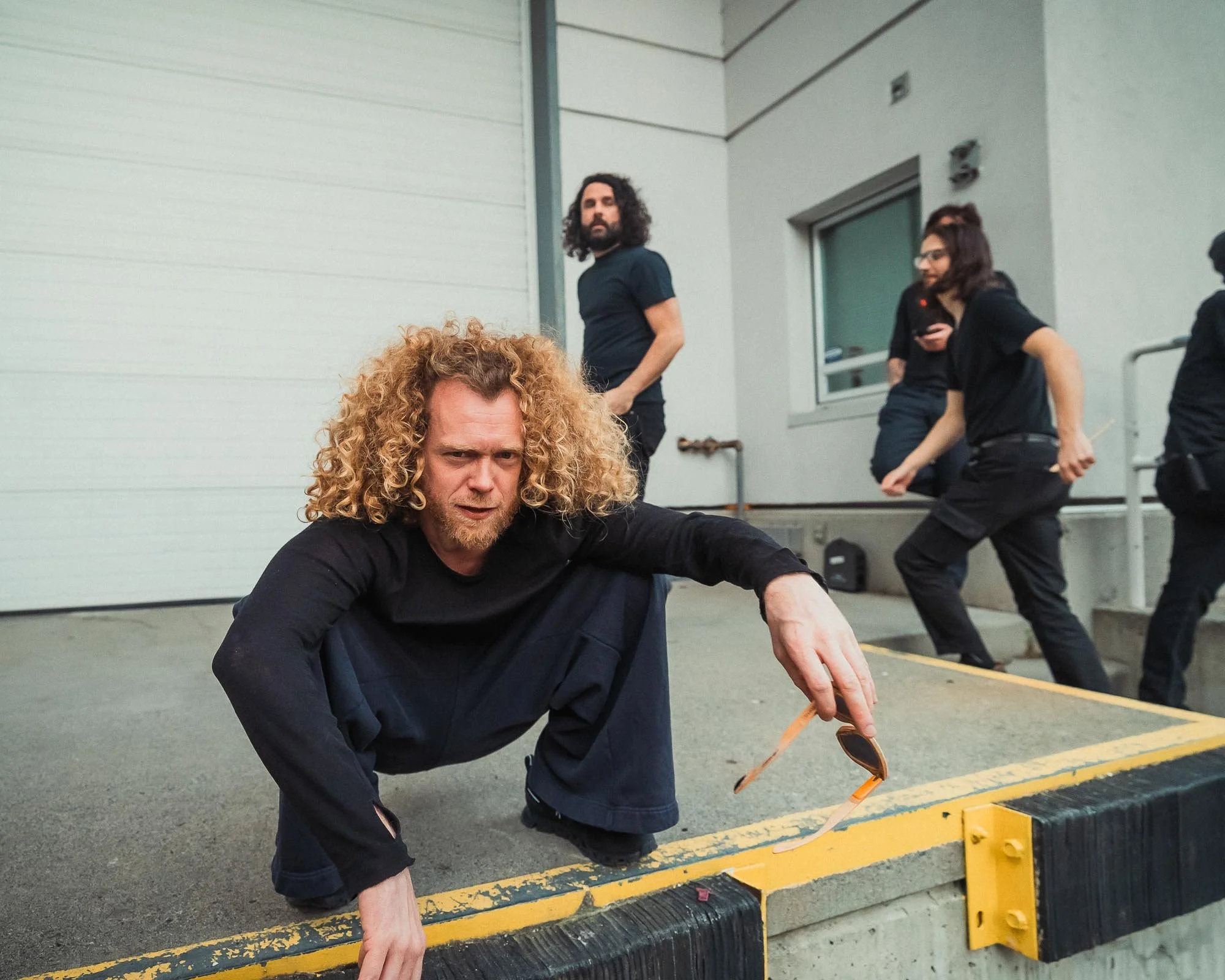 Glenn with curly hair and a beard crouches on the ground near a parking lot barrier, holding a wooden stick with sunglasses hanging from it, with several people standing in the background near a building.