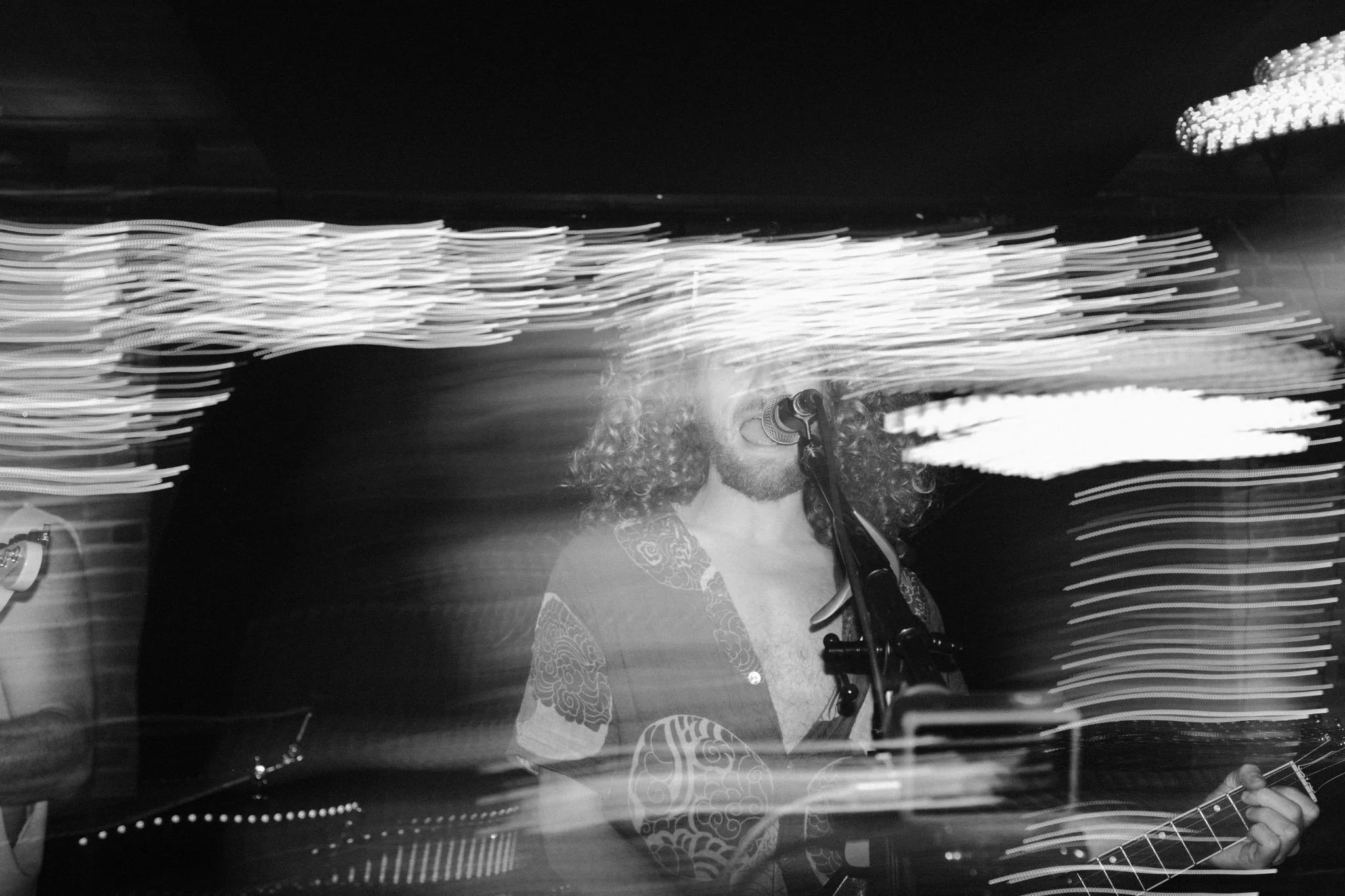 A person with curly hair singing into a microphone and playing an electric guitar, surrounded by streaks of light in a dark setting.