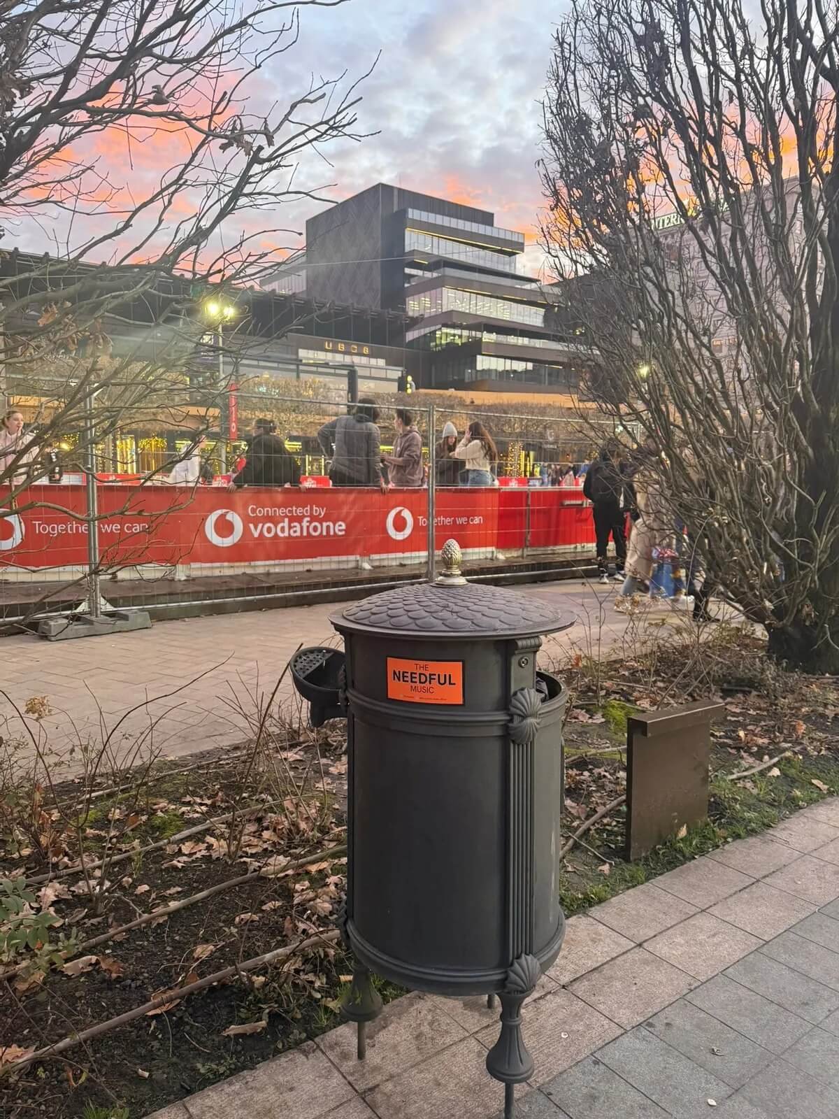 An outdoor scene at dusk showing a street with pedestrians, a modern building in the background, and leafless trees. There is a black trash can with an orange sticker that says 'The Needful Music' in the foreground.