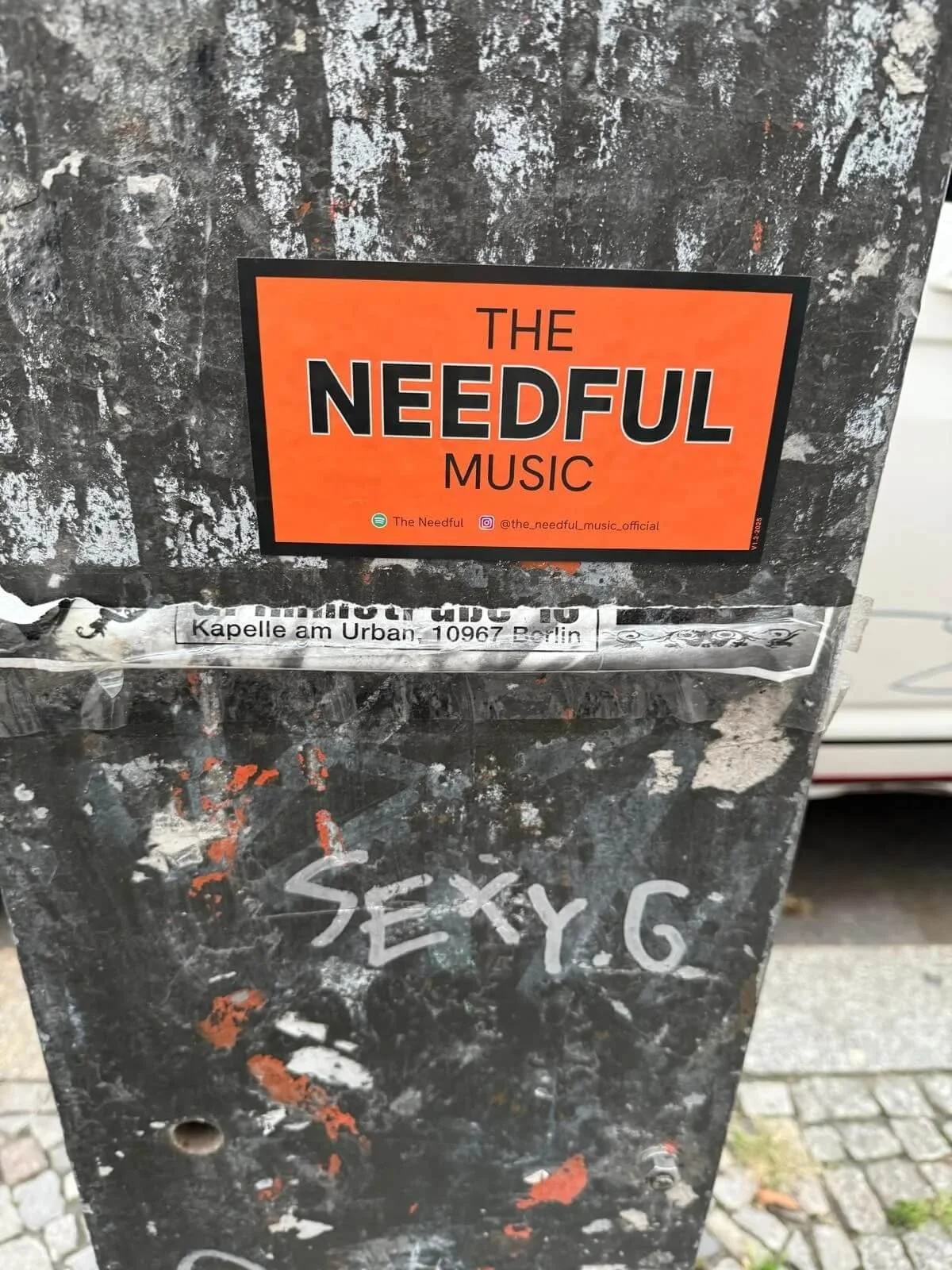 Close-up of a weathered, black, graffiti-covered pole with a bright orange sticker that reads 'The Needleful Music' and social media icons at the bottom. Graffiti with the text 'SEXY G' is visible on the pole.