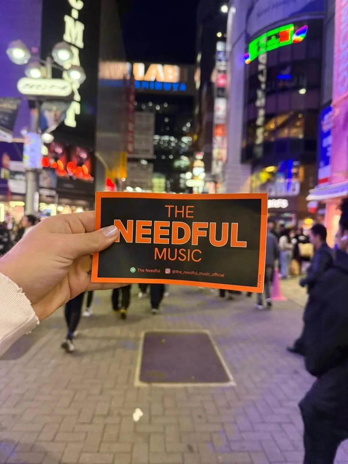 A hand holding a promotional card for The Needful Music in a busy city street at night, with neon signs and people walking in the background.