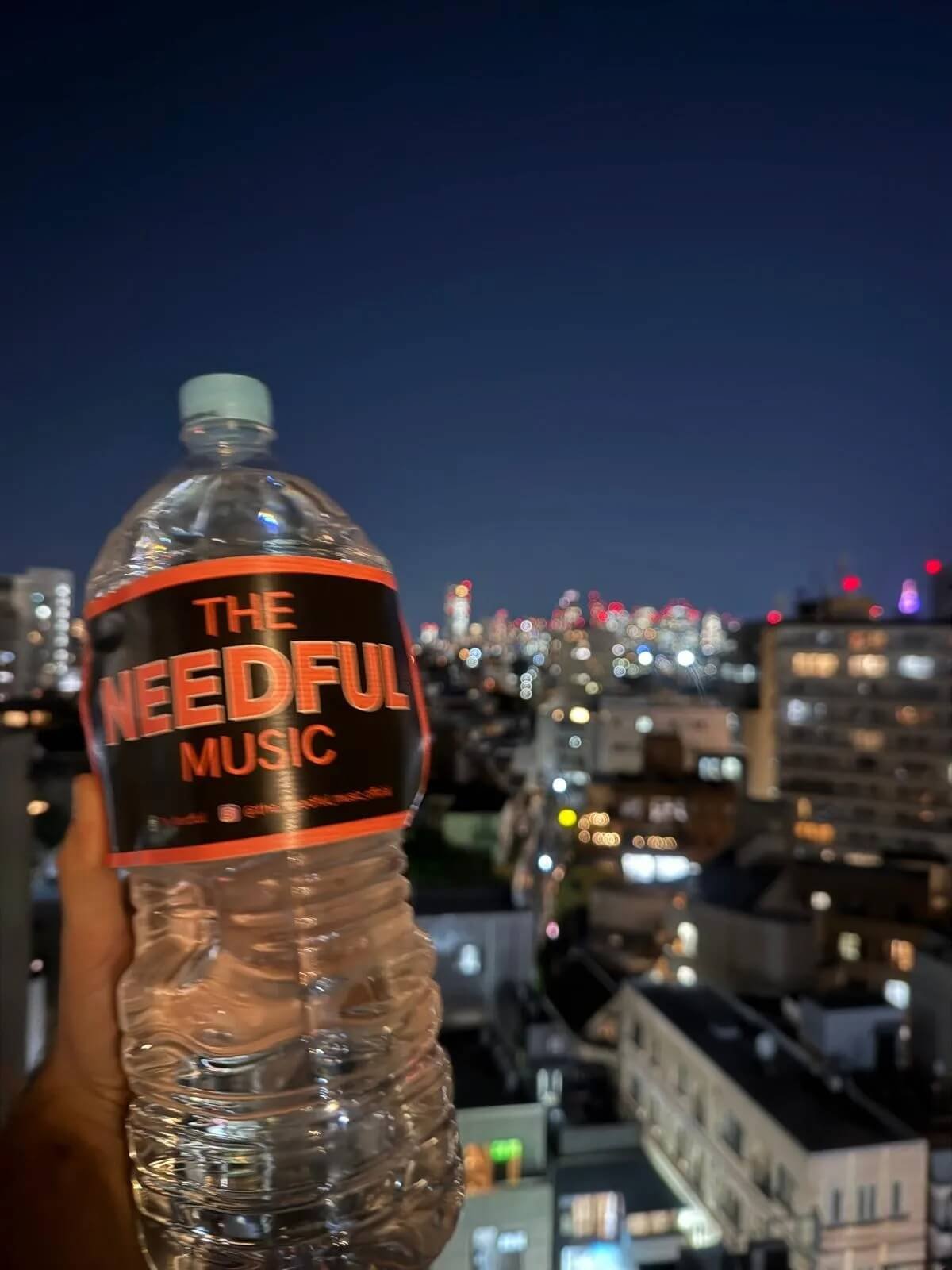A hand holding a clear plastic water bottle with a black and red label that reads 'The Needful Music,' against a blurred city skyline at night with illuminated buildings and a dark sky.