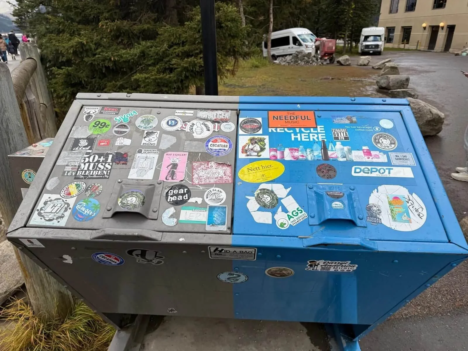 A metal recycling bin with a blue section on the right, covered in stickers and labels, located outdoors near a sidewalk with trees, rocks, and parked vans in the background.