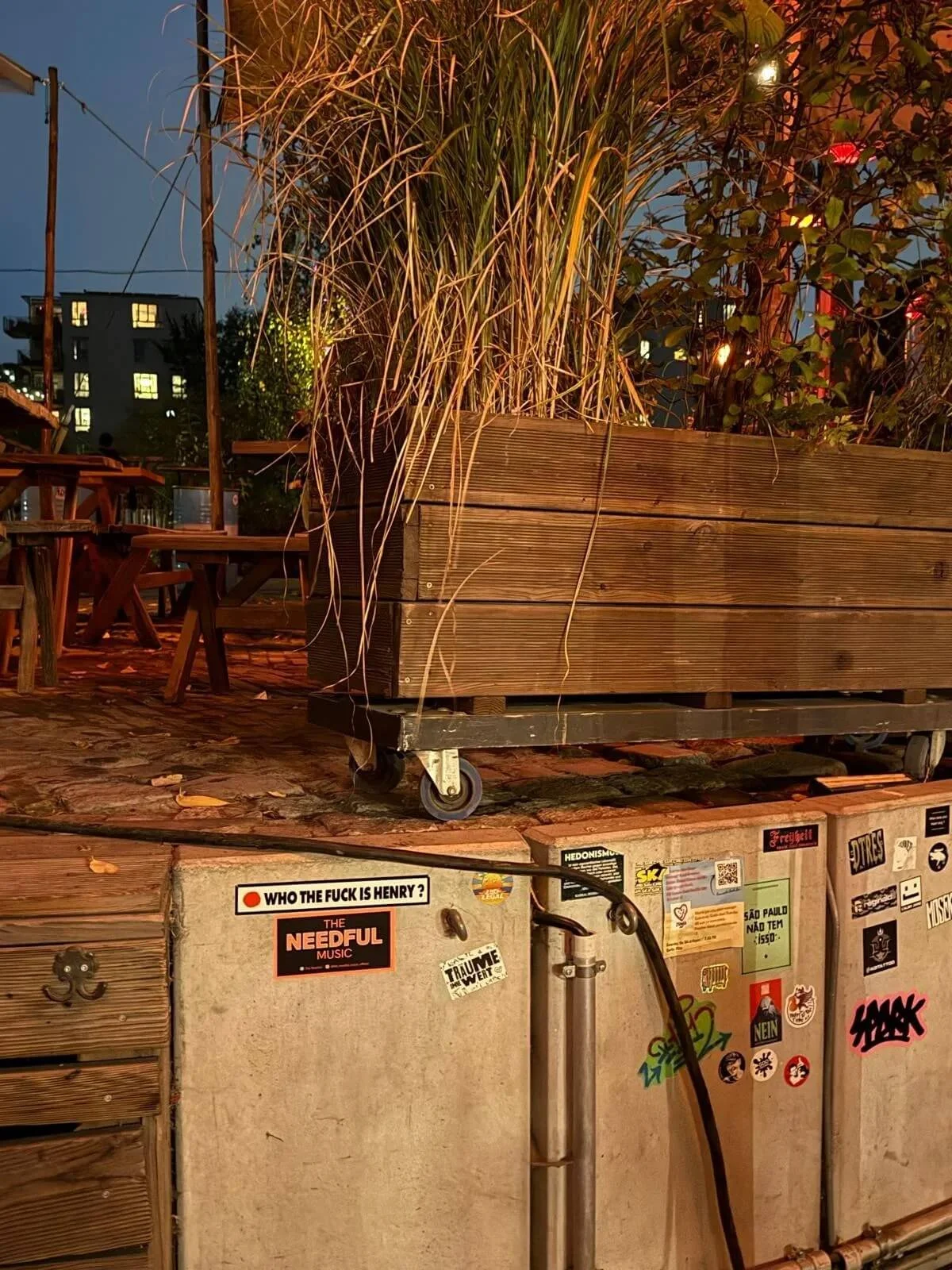 Outdoor patio scene with wooden planters filled with plants, patio furniture, evening sky, and a wall with various stickers, posters, and signs.