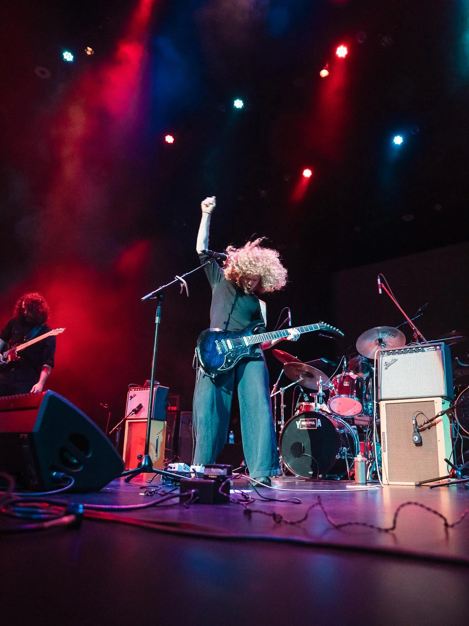A man with curly blonde hair playing an electric guitar on stage, with colourful stage lights overhead, and a drummer and other musicians in the background.