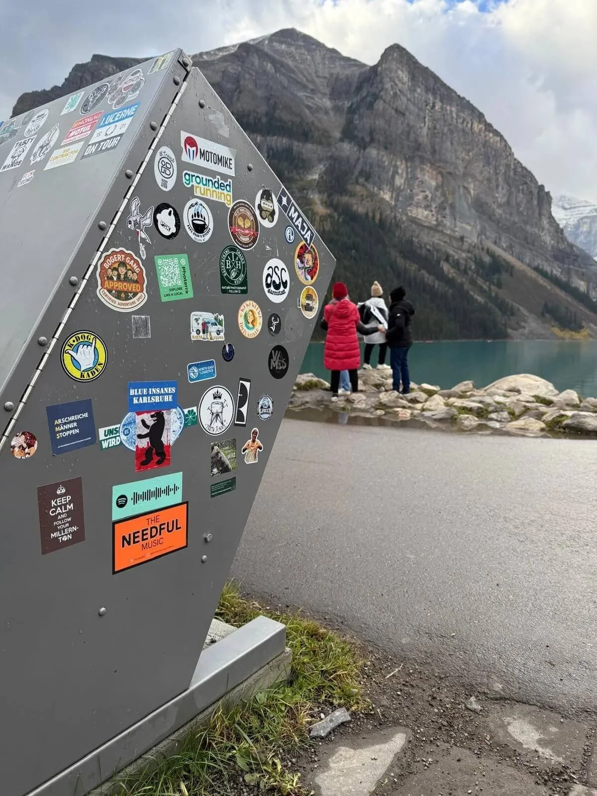 A gray metal surface covered with numerous stickers, set outdoors near a lake with mountains in the background. Three people in winter clothing are standing near the lake, facing the water, with one person in a red coat, two in black and white coats.