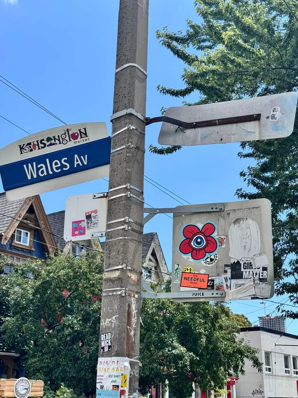 Street pole with a blue street sign labeled 'Wales Ave', surrounded by various stickers and flyers. Behind the pole, there are trees and houses with detailed architecture, and the sky is clear with some power lines overhead.