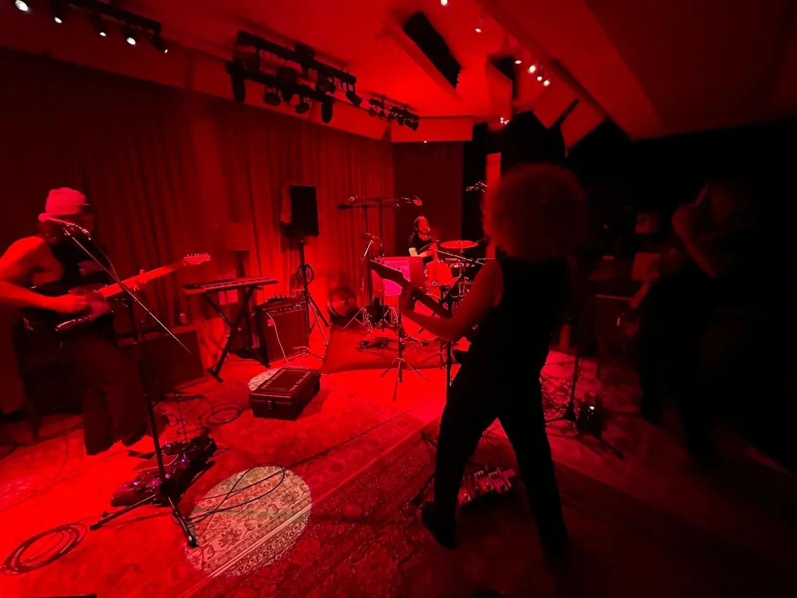 The Needful band performing on a stage with red lighting, featuring a guitarist, a drummer, and another musician, with a red curtain backdrop.