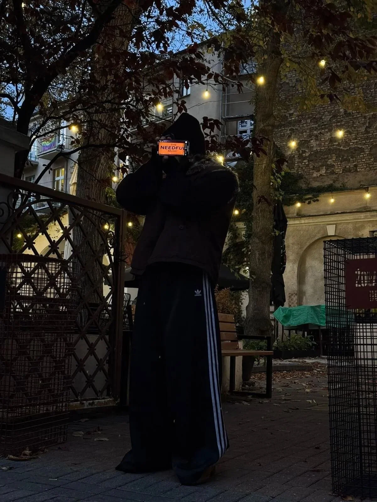 Person standing outdoors at night, wearing a black jacket, black pants with white stripes, and a black hat, holding a phone to their ear. The phone's screen displays the word 'NEEDFUL'. The background includes a tree with string lights, a brick build