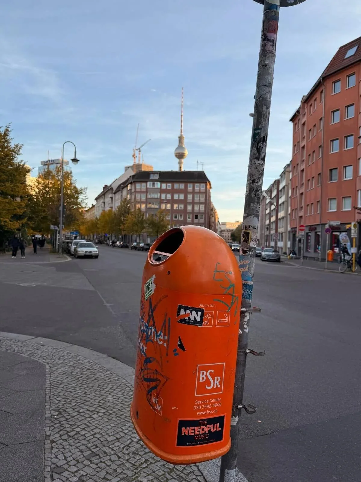 An orange trash can with stickers on a street corner in Berlin, Germany, with the Berlin TV Tower in the background. The street has a few cars and pedestrians, with buildings on either side.