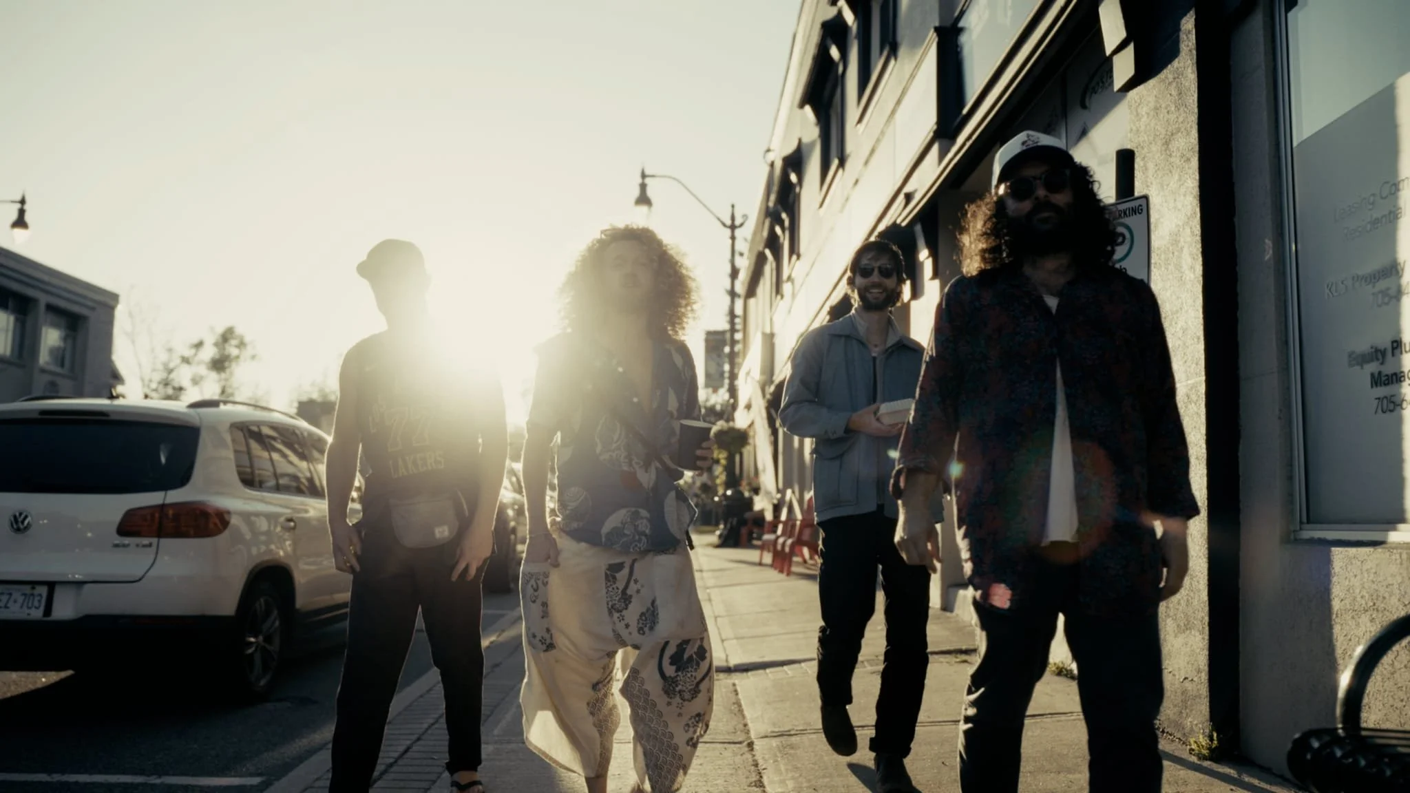 The Needful band members walking on a city sidewalk during sunset, with cars parked along the street and buildings in the background.