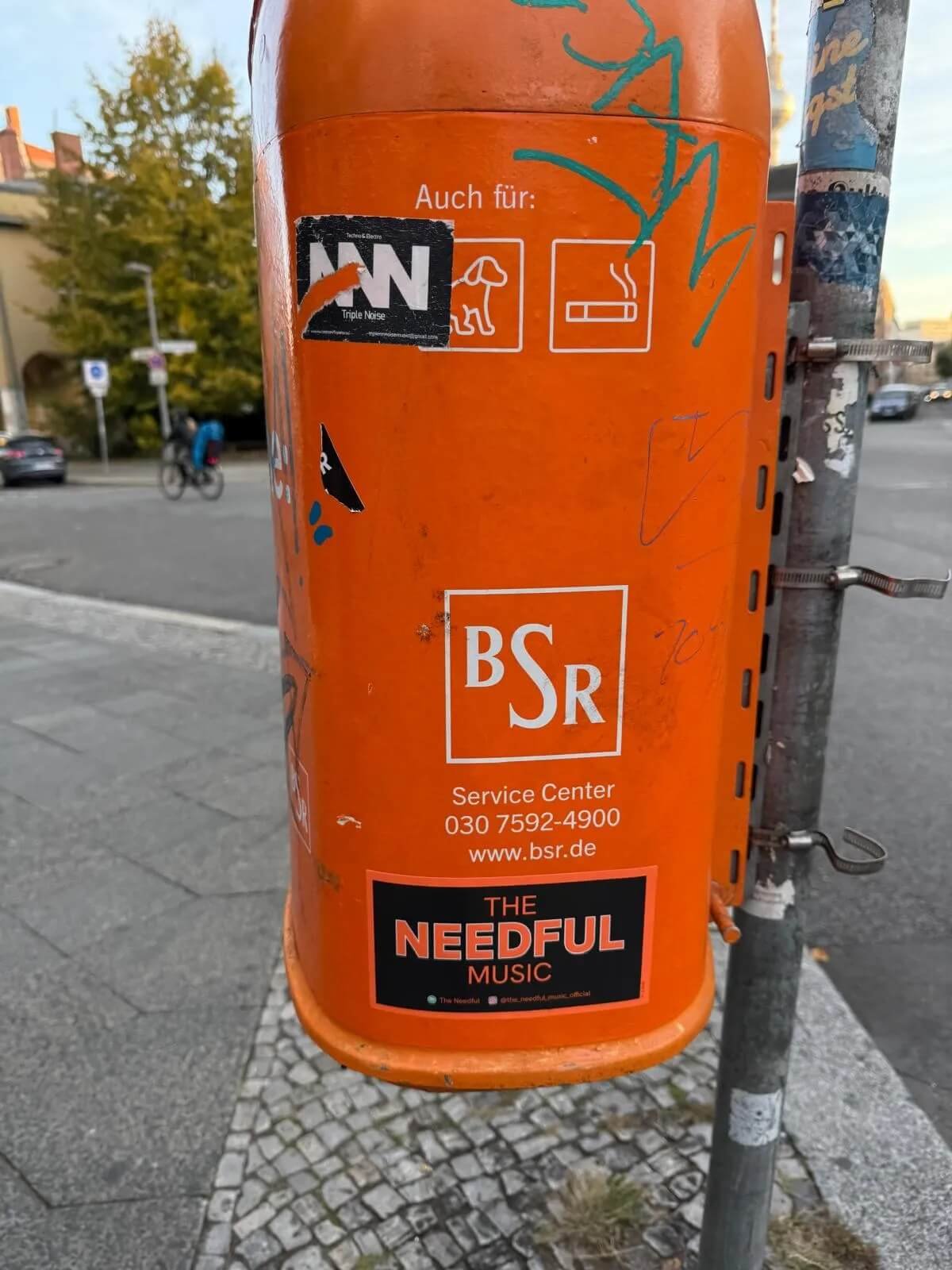 Orange mailbox with stickers and graffiti, including a sticker for 'The Needful Music,' standing on a sidewalk with a street and trees in the background.