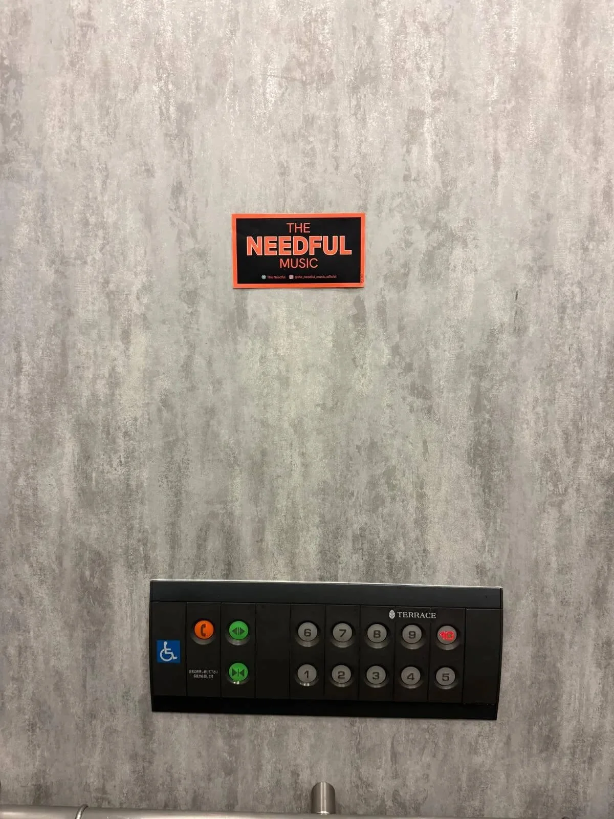 Elevator control panel with numbered buttons, call buttons, and accessibility symbol, mounted on a gray wall with a sign reading 'The Needful Music' above it.
