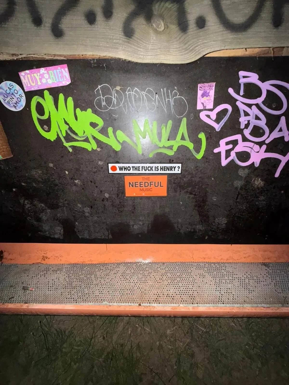 A graffiti-covered metal surface with various stickers on it, including a pink sticker that says 'MUY BIEN,' a blue sticker that says 'LUCIAS PINTO AMOR,' a purple sticker with unreadable text, and two stickers with hearts and words in Spanish. There