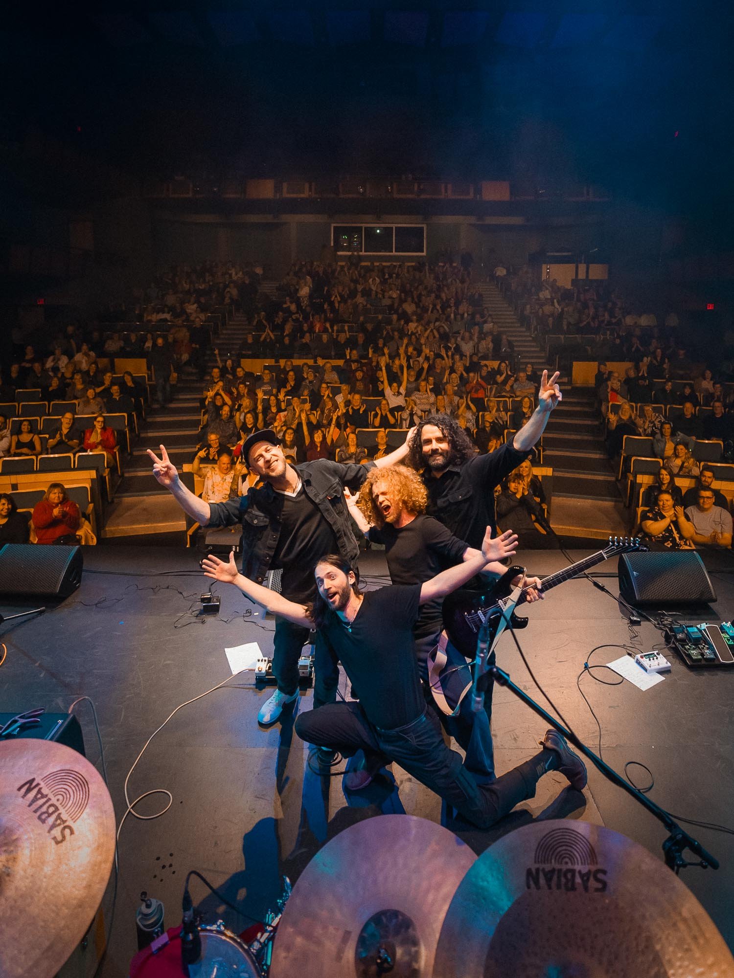 View from the stage of a concert with The Neddful drummer's cymbals in the foreground and four musicians smiling and posing for the camera with an audience clapping and cheering in the background.