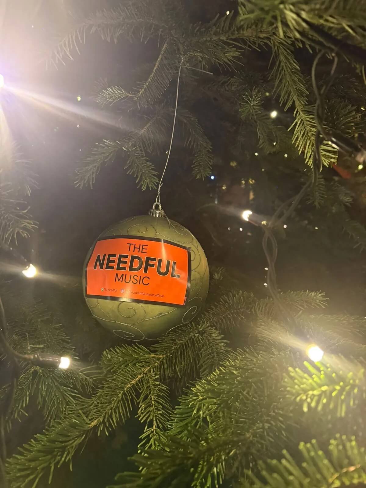 A green Christmas ornament with swirl designs hangs on a decorated Christmas tree, with a label reading 'THE NEEDFUL MUSIC' attached to it. The tree is decorated with small white lights, and the background includes some blurred lights and branches.