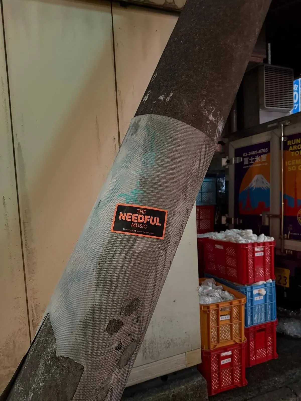 A gray metal pipe with a sticker that says "The Needful Music" is leaning against a wall. In the background, there are red and orange plastic crates filled with plastic bags, and an industrial setting with signs and equipment.