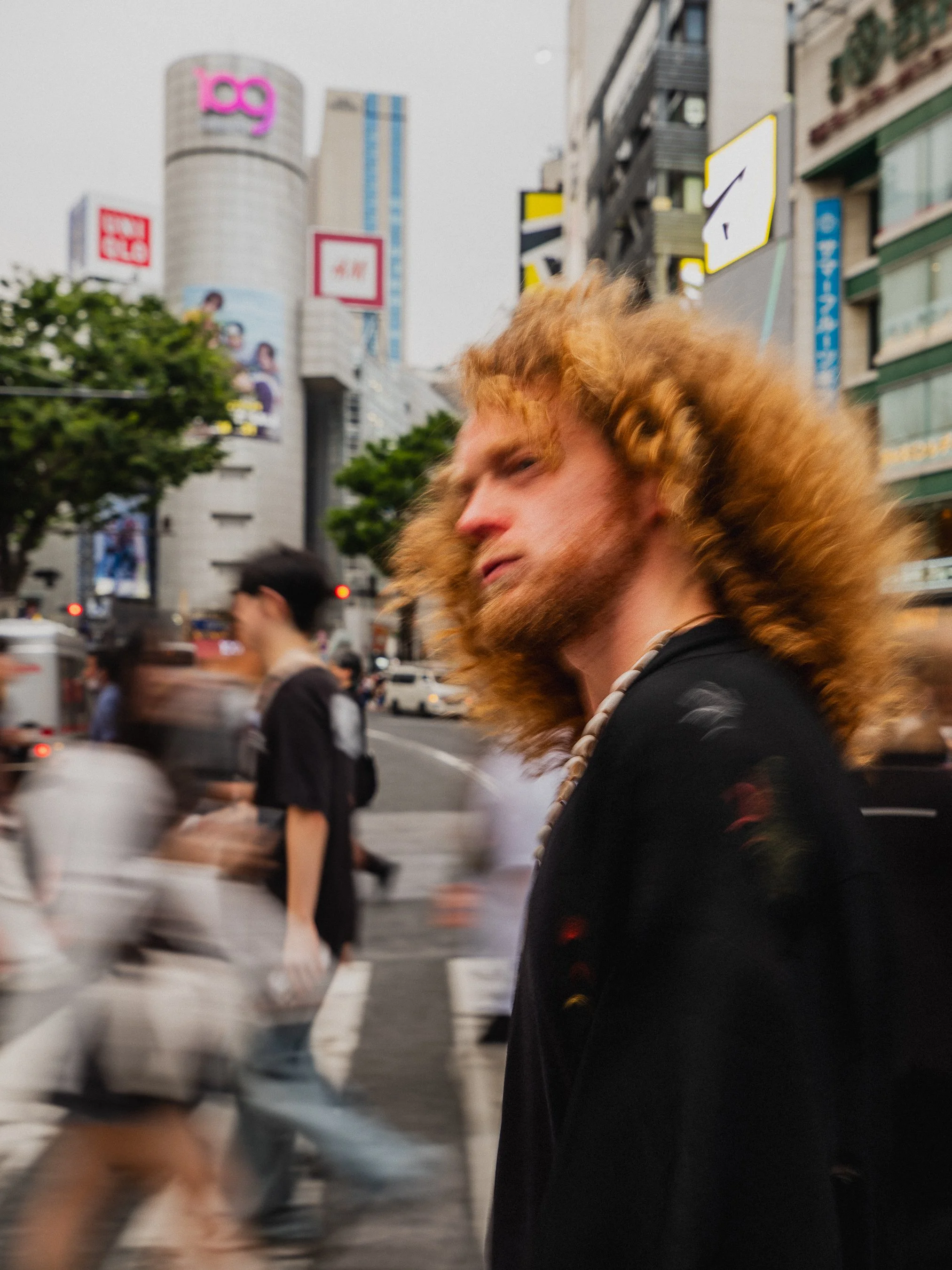 singer for The Needful, with curly red hair and a beard, wearing a black jacket and a shell necklace, walking on a busy city street with blurred pedestrians and tall buildings, including a Disney store, in the background.