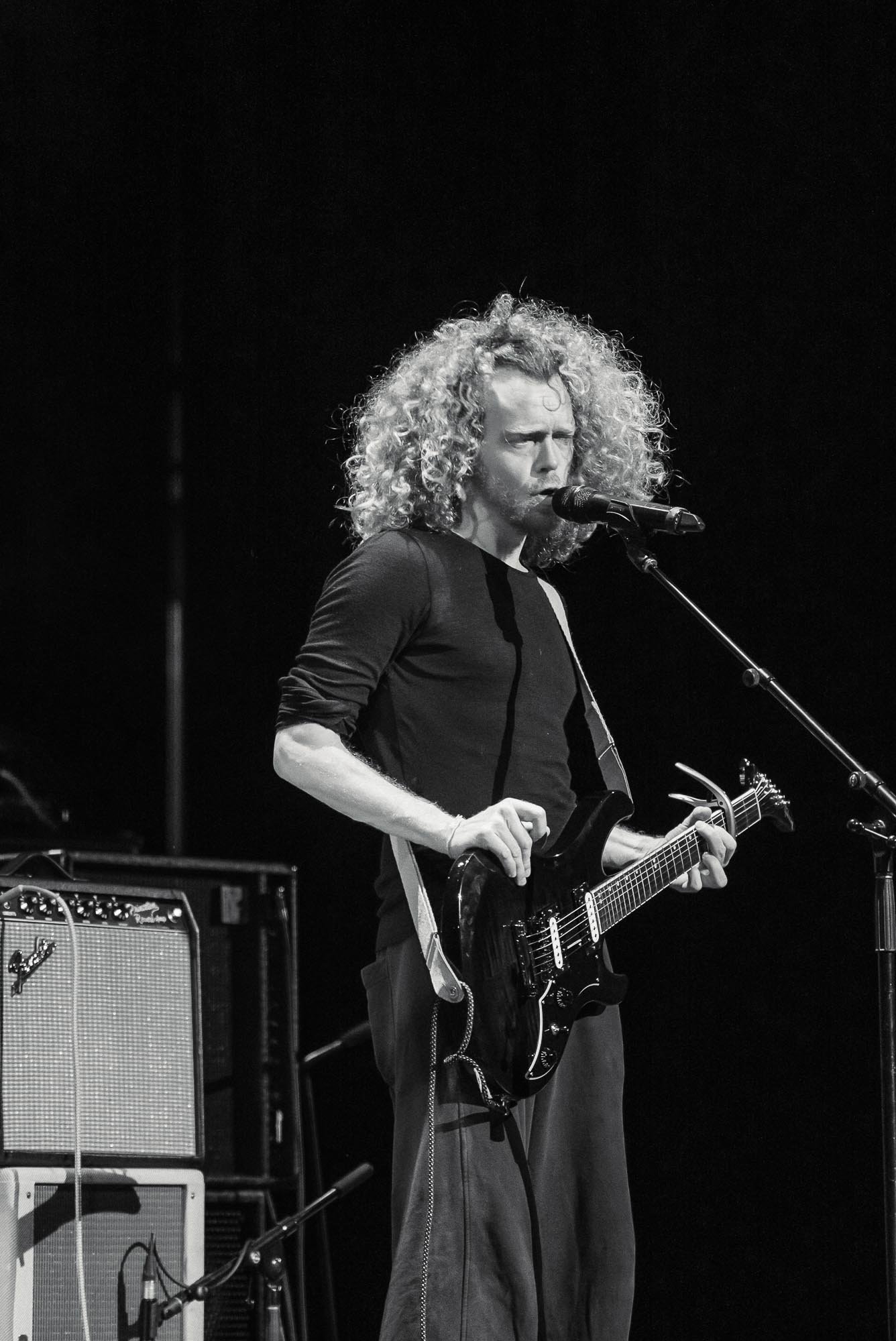 A man with curly blonde hair is performing on stage, singing into a microphone and playing an electric guitar. He is wearing a dark long-sleeve shirt and loose pants. There are musical equipment and amplifiers visible in the background.
