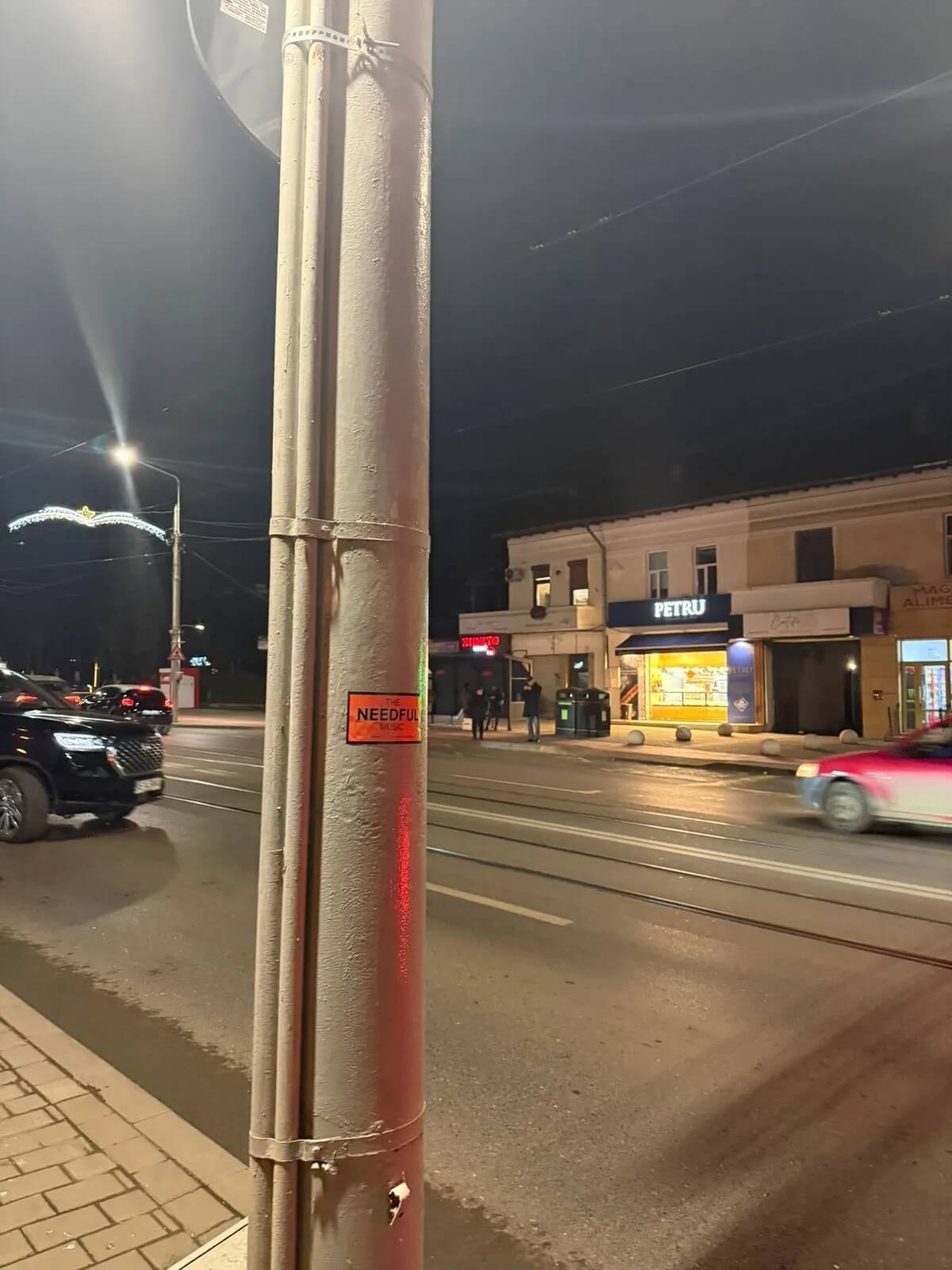 Night street scene with cars driving on the road, a lamp post, and a shop named PETRU with people nearby.