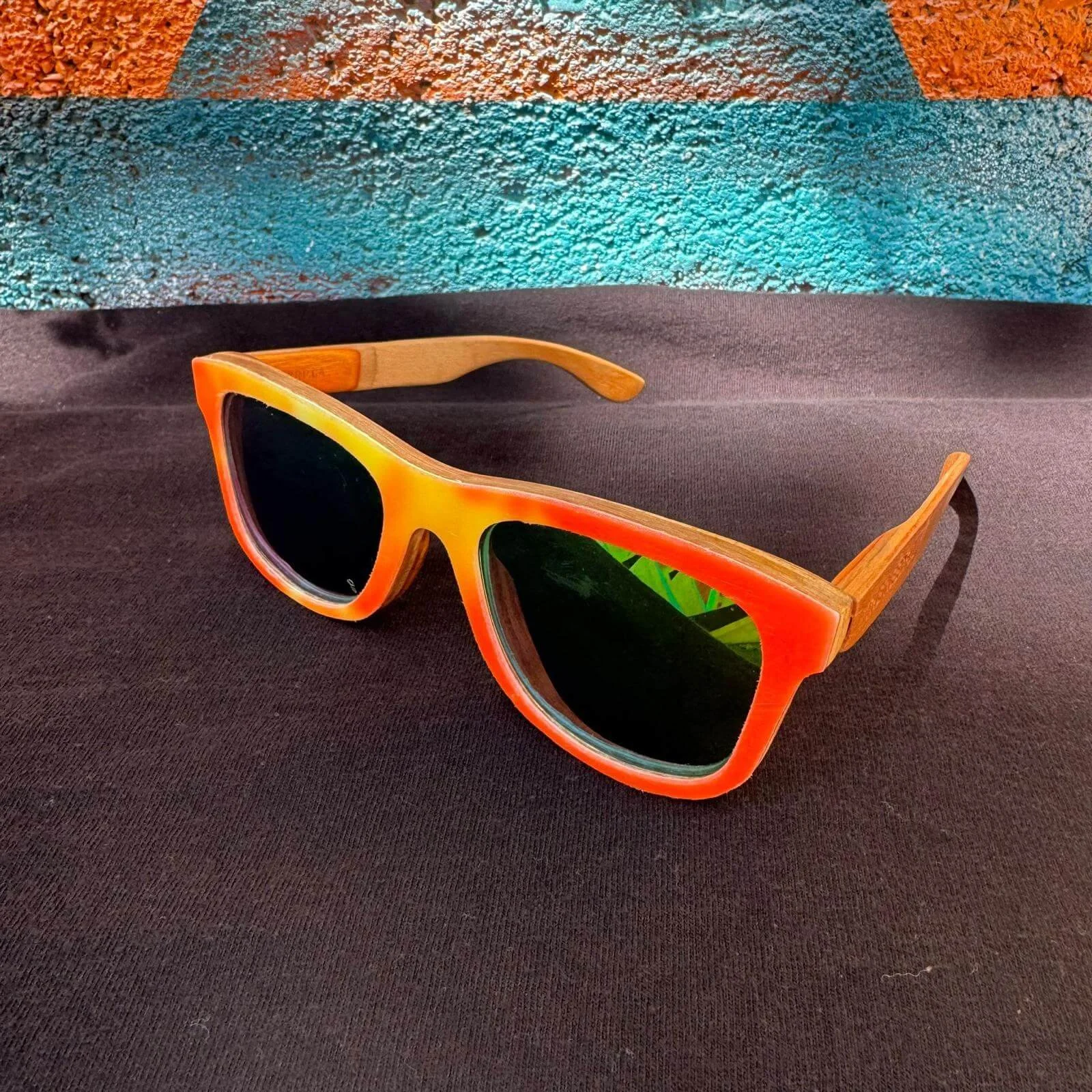 Colorful wayfarer sunglasses with orange and yellow frame and dark lenses resting on a black surface. Background features a textured wall with orange, turquoise, and earthy tones.
