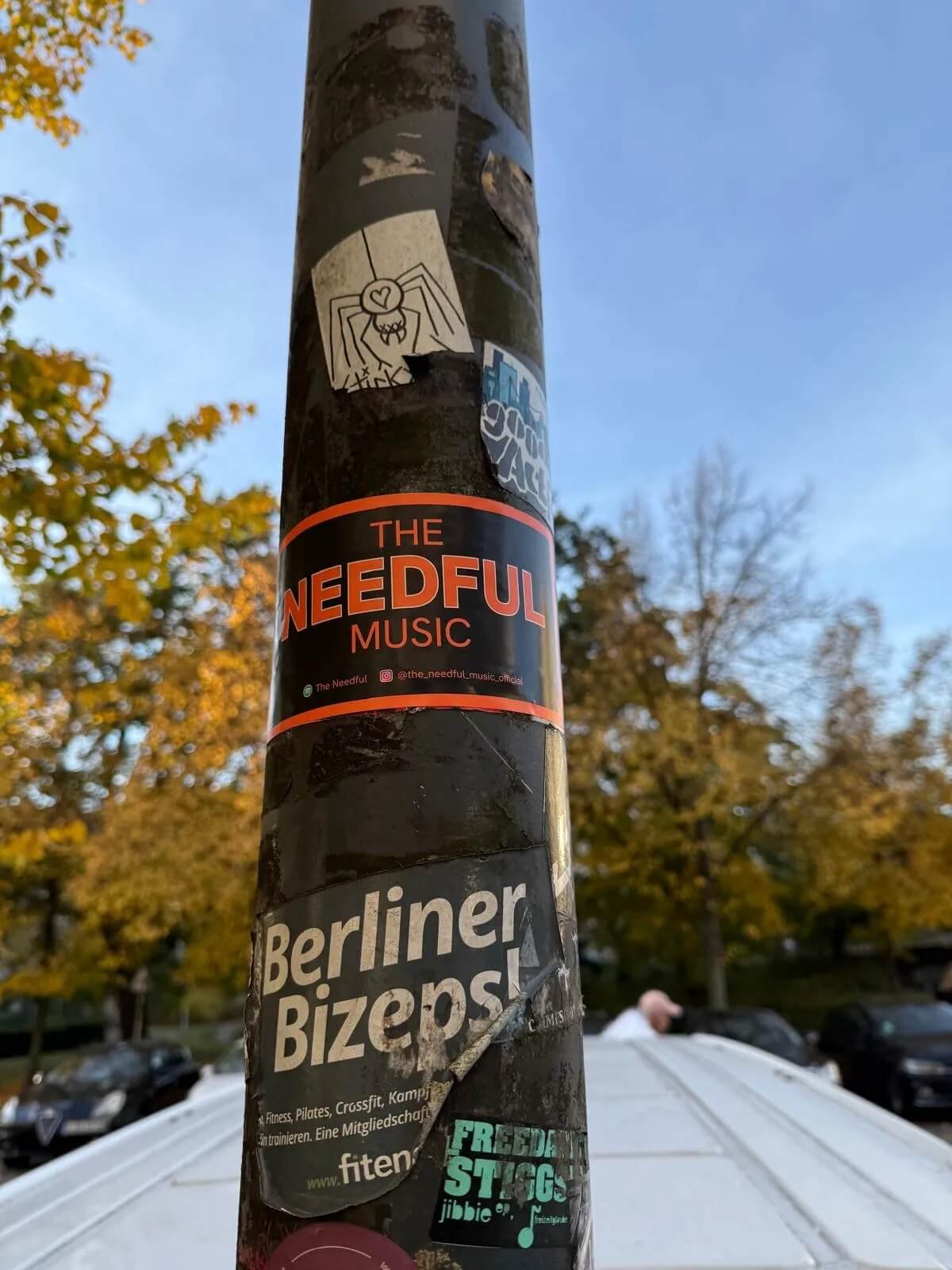 A black pole covered with various stickers, including a sticker for 'The Needful Music', a sticker labeled 'Berliner Bizeps', and a few smaller, less readable stickers. The background shows a clear sky with some autumn-colored trees and part of a car