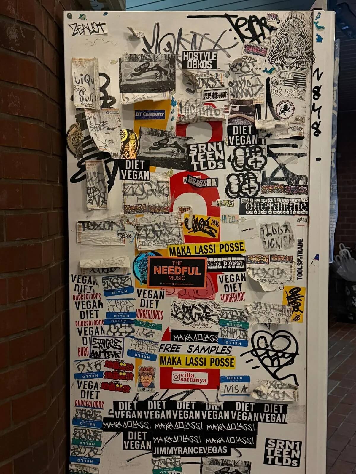 A white bulletin board covered with overlapping stickers, graffiti tags, and small posters. The stickers include messages like "Vegan Diet," "Maka Lassi Posse," 
