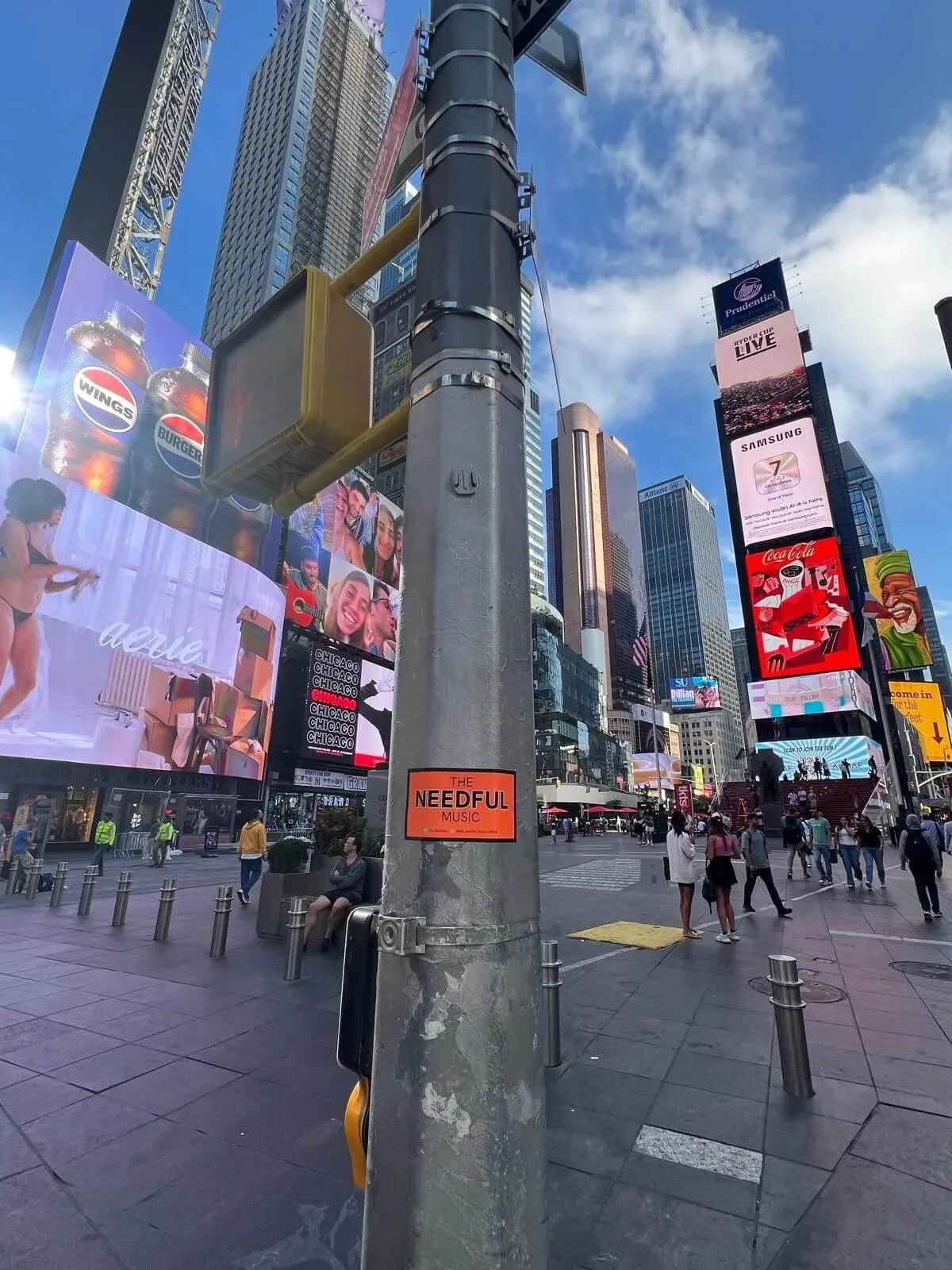 Times Square in New York City with numerous digital billboards, tall skyscrapers, and pedestrians. There is a streetlight in the foreground with a small orange sticker that says "The Needful Music."