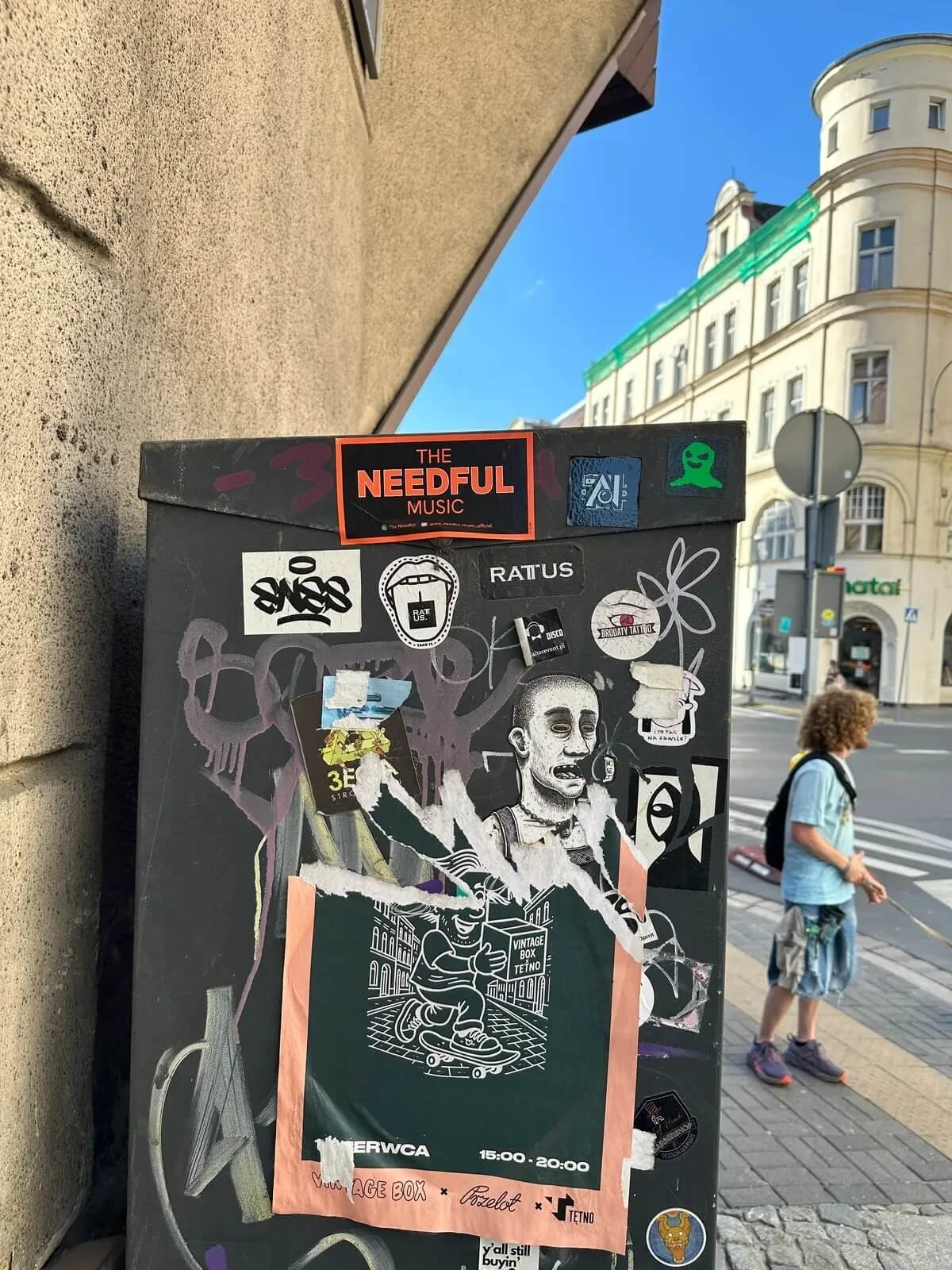 A black outdoor poster board covered with various stickers and torn paper posters, placed on a street corner against a wall. In the background, a person with curly hair wearing a light blue shirt and shorts is walking past, with a city street and bui