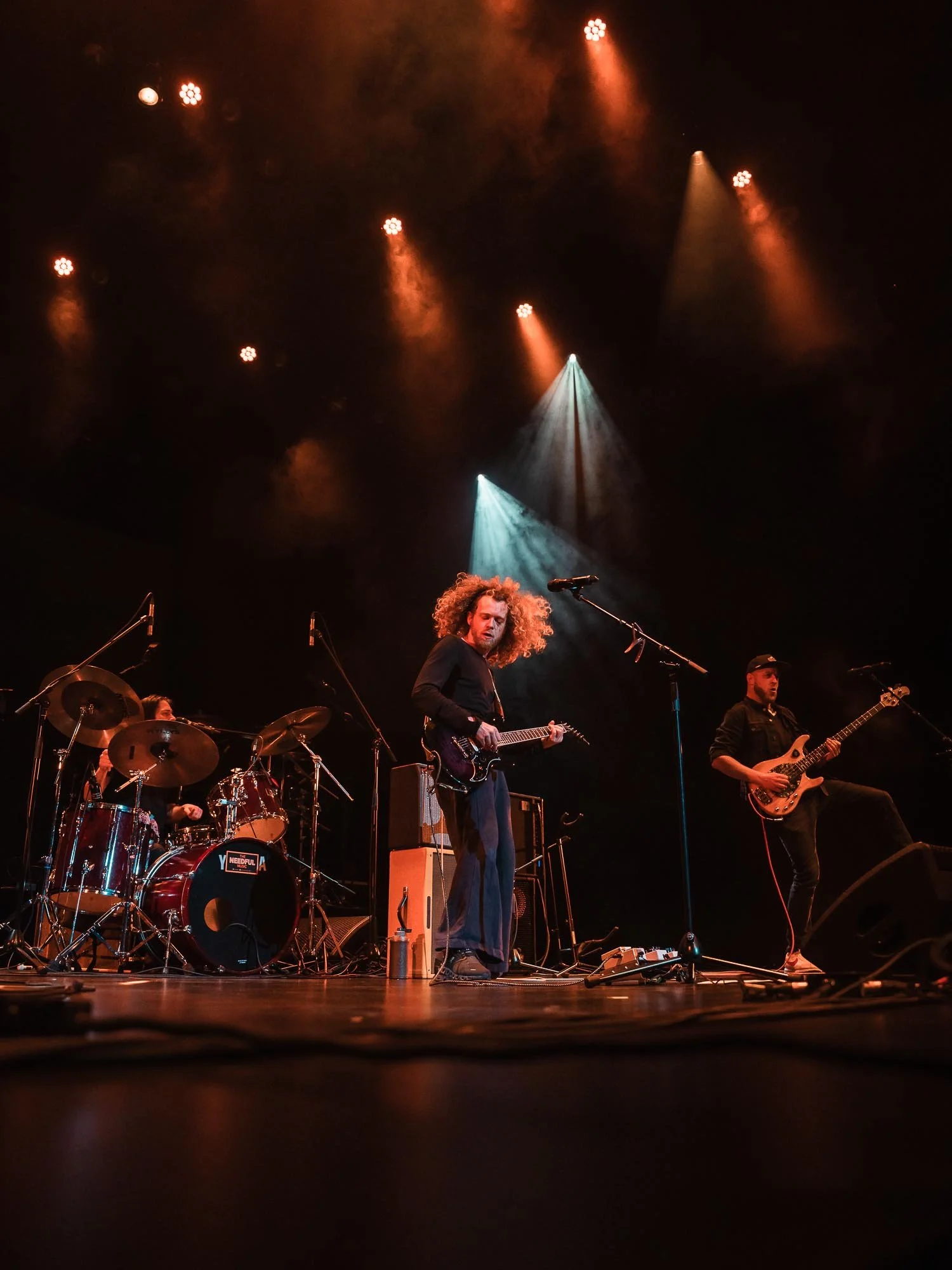 The Needful alt-indie musicians performing on stage under orange and blue stage lights, with a drummer, guitarist, and bassist visible.